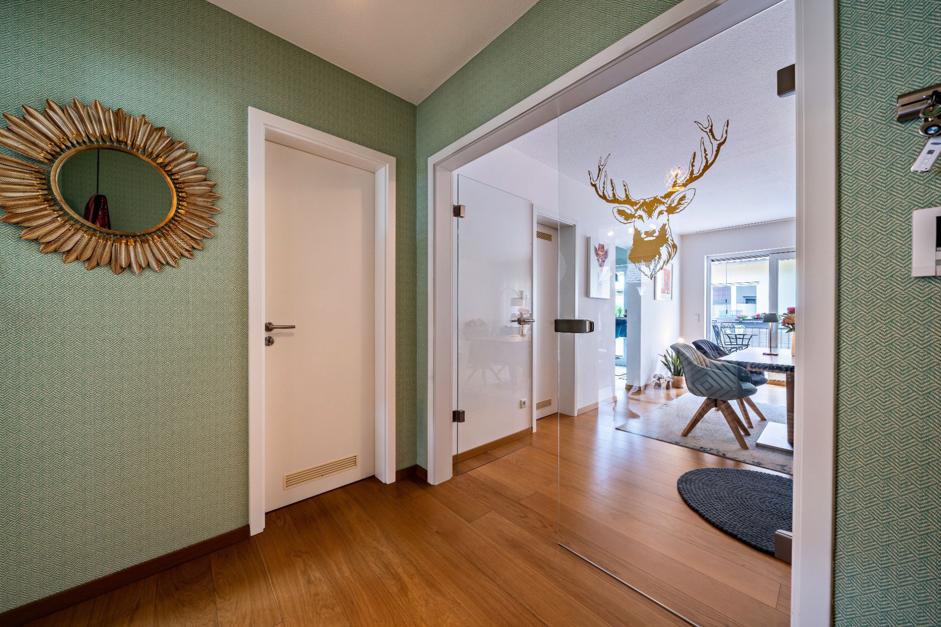 Flur mit Holzboden, grüner Wand, Spiegel und Glastür mit Blick ins Wohnzimmer.