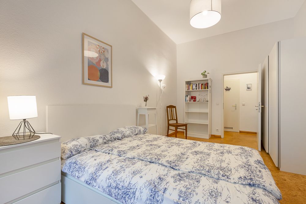 Helles Schlafzimmer mit Doppelbett, Nachttisch, Kommode, Regal, Stuhl und Wandbild.