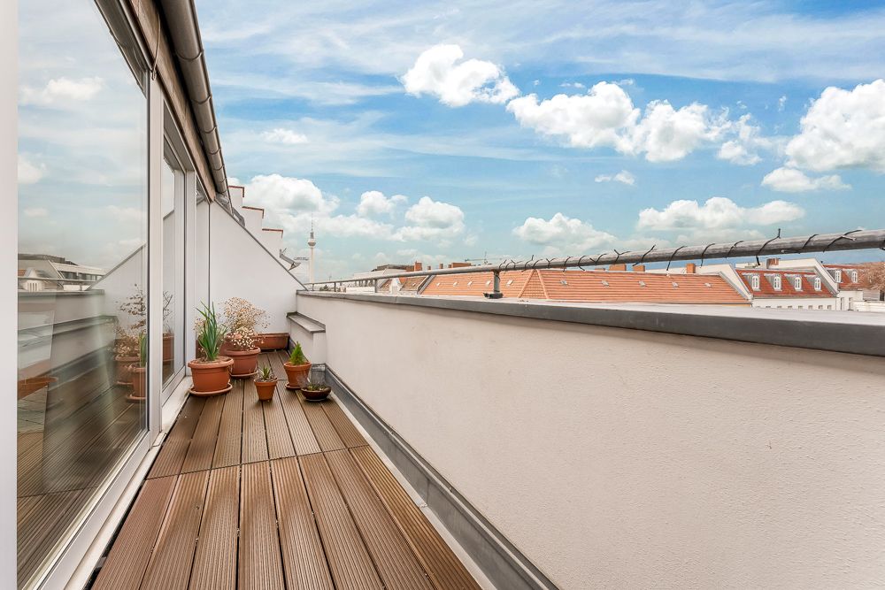 Schmaler Balkon mit Holzboden, Pflanzentöpfen und Blick auf Dächer unter blauem Himmel.