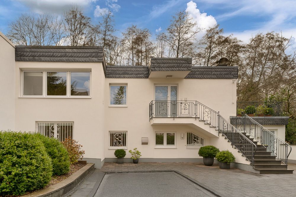 Moderne zweistöckige Wohnhausfassade mit Außentreppe, Balkon und Pflanzen vor dem Haus.