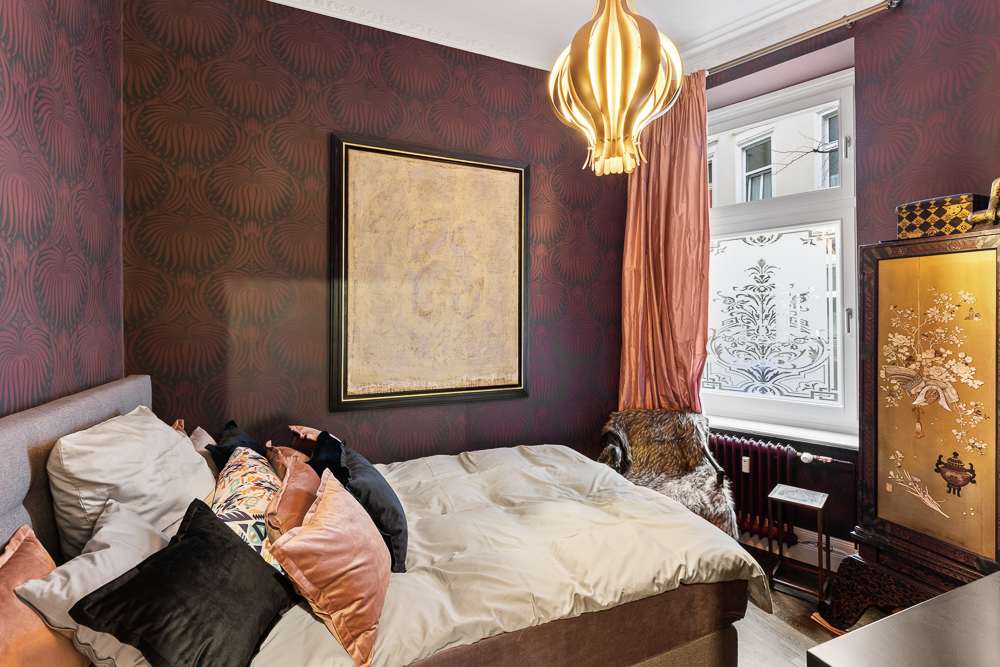 Kleines Schlafzimmer mit Doppelbett, gemusterten Kissen, goldener Lampe und Fenster mit Vorhang.