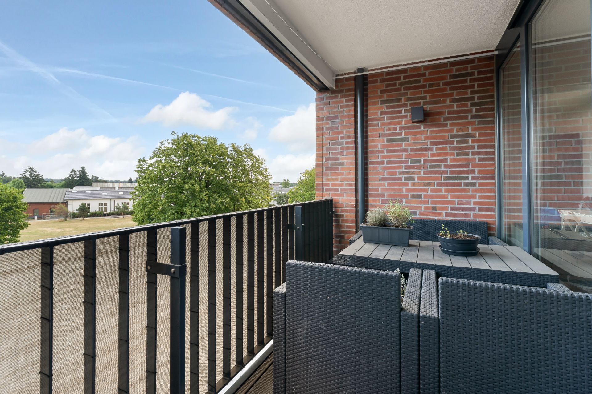 Balkon mit Gartentisch, zwei Stühlen und Blick auf Bäume sowie Gebäude im Hintergrund.