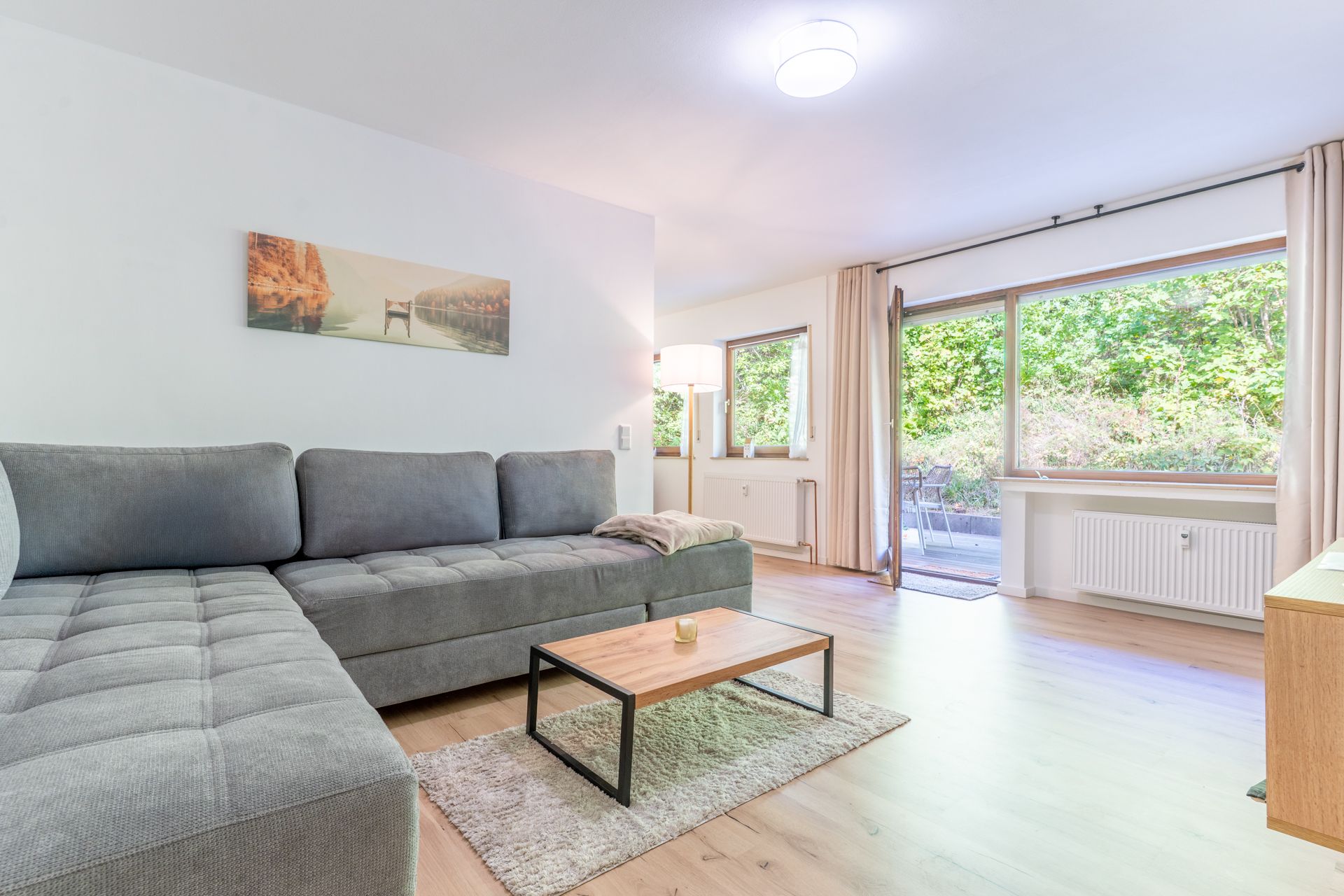 Helles Wohnzimmer mit grauem Ecksofa, kleinem Holztisch und großem Fenster mit Blick ins Grüne.