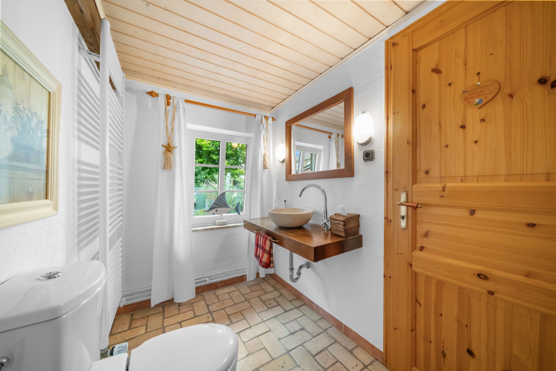 Helles Bad mit Holzdecke, großem Spiegel, Waschbecken, Toilette und Fenster mit Blick ins Grüne.
