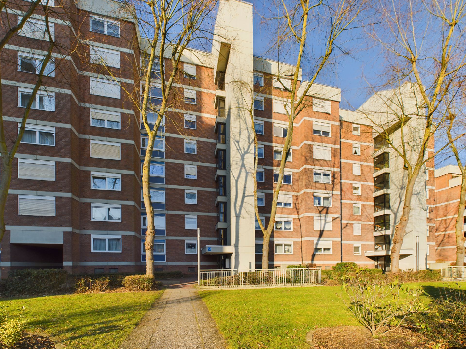 Mehrstöckiges Wohnhaus mit roten Backsteinwänden und vielen Fenstern, davor Rasen und Bäume.