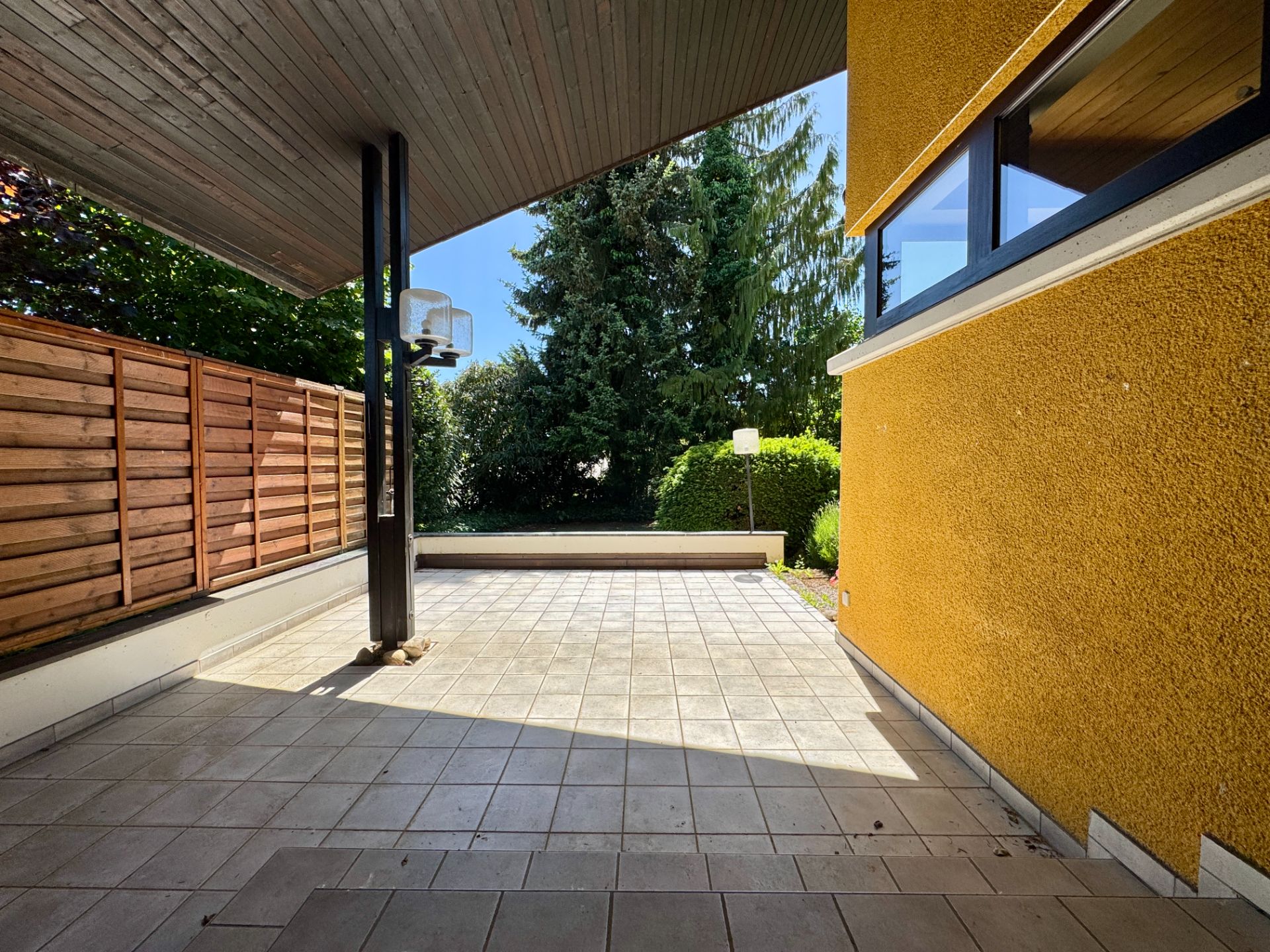 Überdachte Terrasse mit Fliesenboden, Holzzaun und Blick auf Garten mit Bäumen.