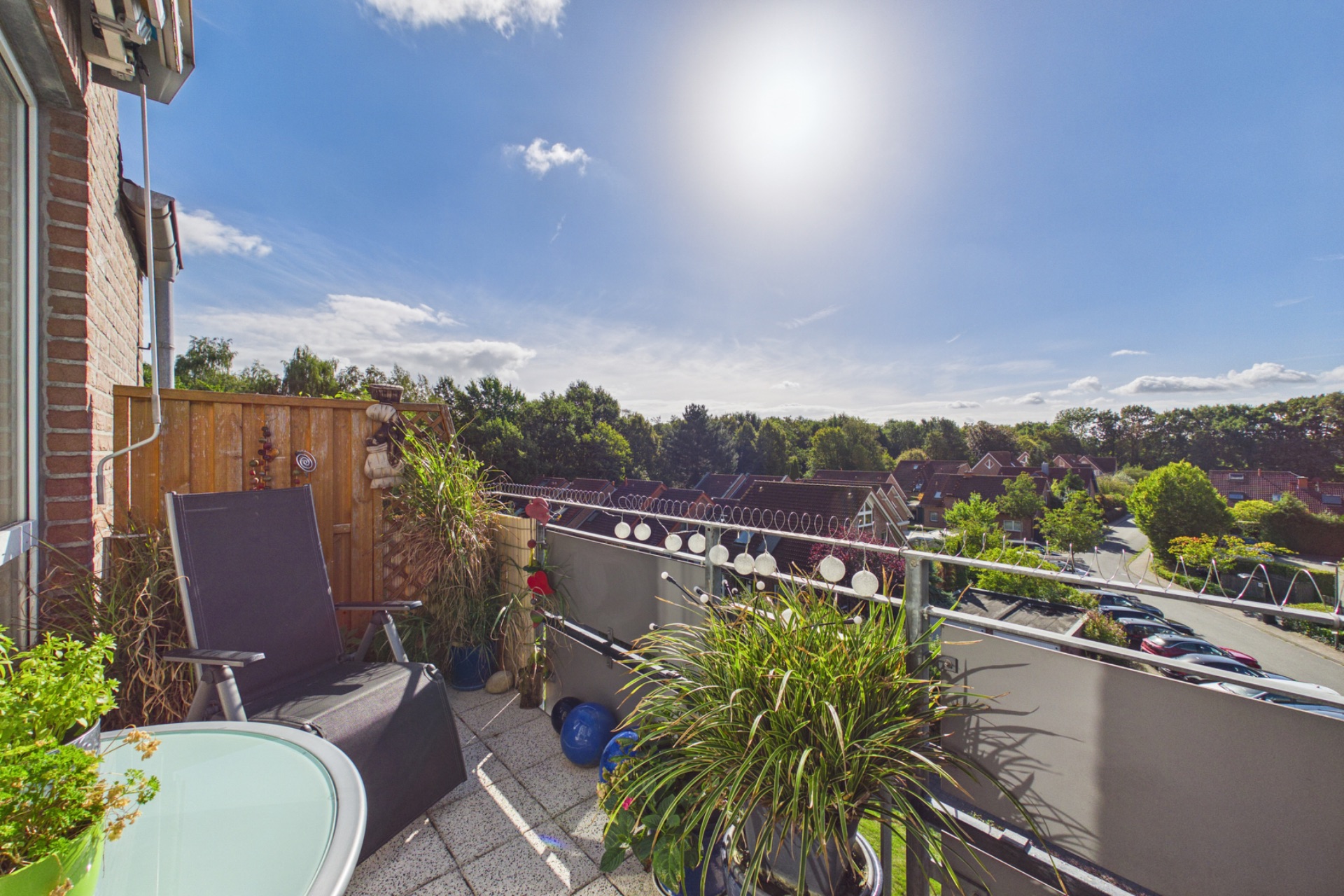 Sonniger Balkon mit Stühlen, Tisch, Pflanzen und Ausblick auf Wohngebiet und Bäume.