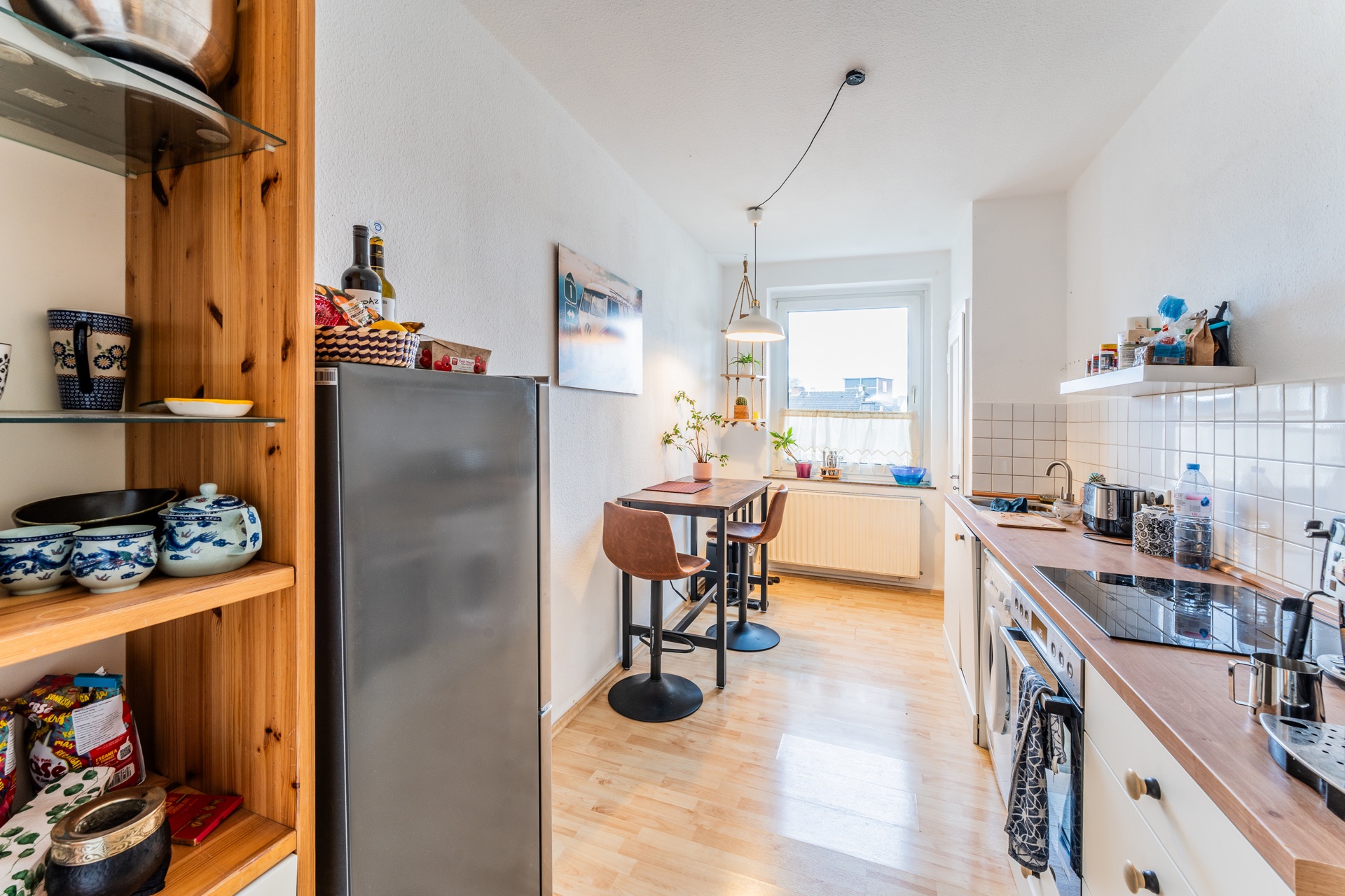 Helle Küche mit Esstisch am Fenster, zwei Stühlen, offenem Regal und modernen Küchengeräten.