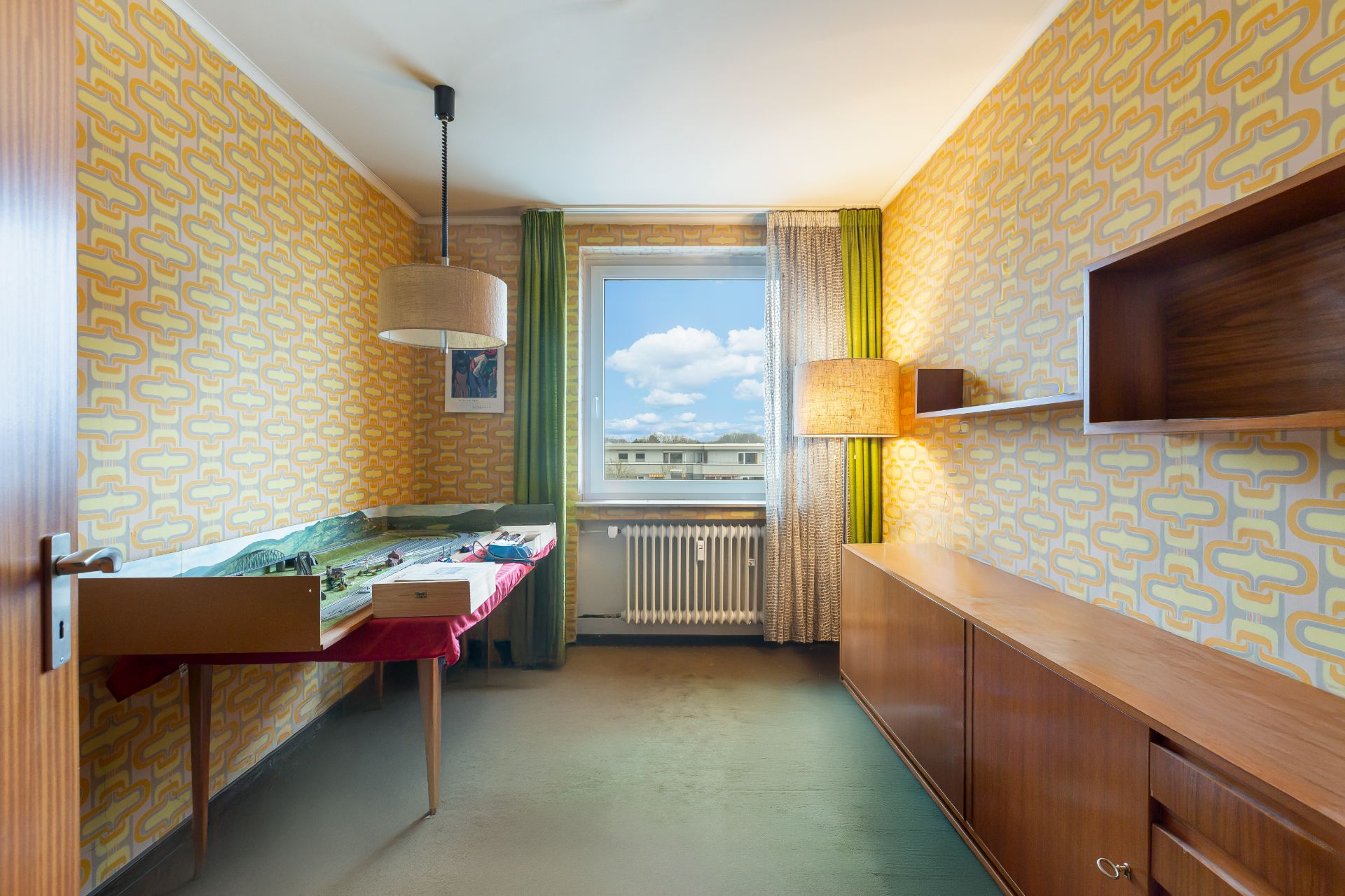 Schmales Zimmer mit Retro-Tapete, Holzmöbeln und Modellanlage auf einem Tisch vor dem Fenster.