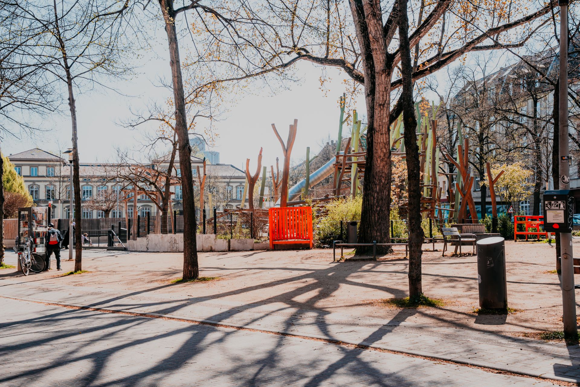 Städtischer Spielplatz mit Bänken und Bäumen, im Hintergrund Wohnhäuser.