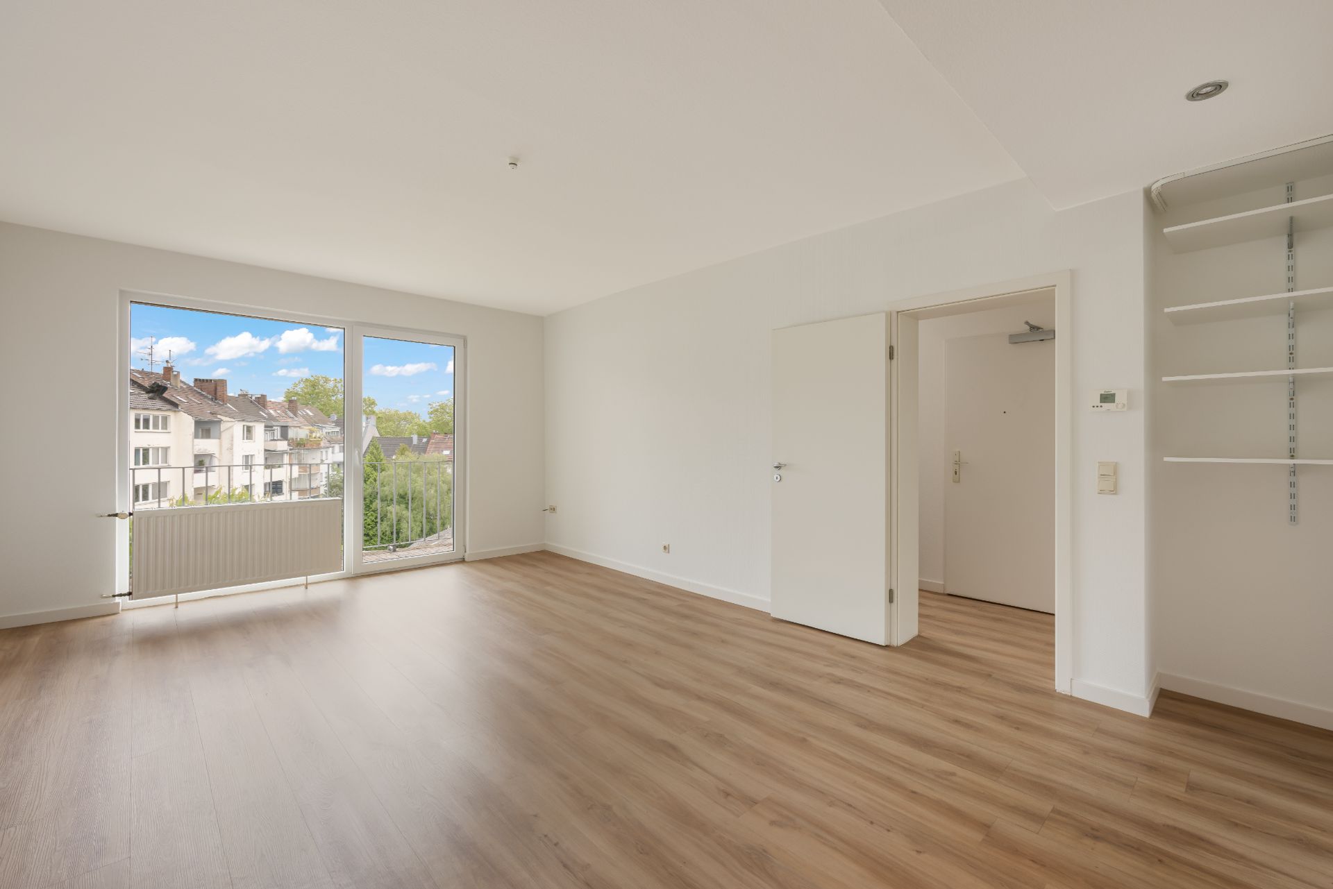 Helles, leeres Zimmer mit Holzfußboden, großem Fenster und offener Tür, Wandregale rechts.