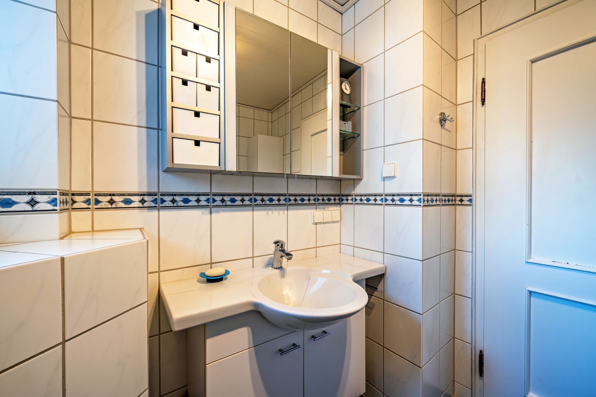 Modernes Badezimmer mit weiß gefliesten Wänden, Waschbecken, Spiegel- und Hängeschrank.