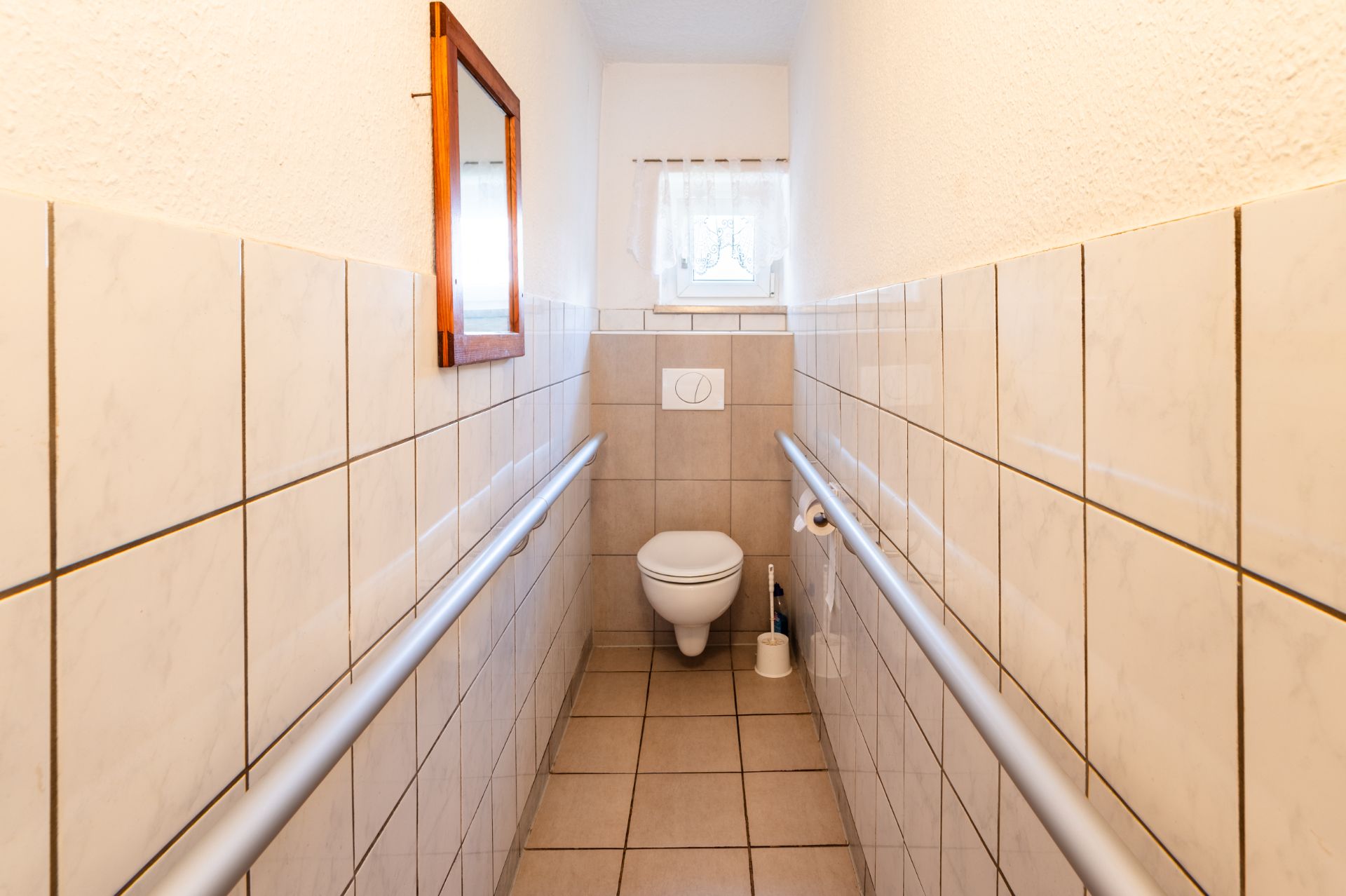 Schmaler gefliester Flur mit Toilette, Haltegriffen an beiden Seiten und kleinem Fenster am Ende.