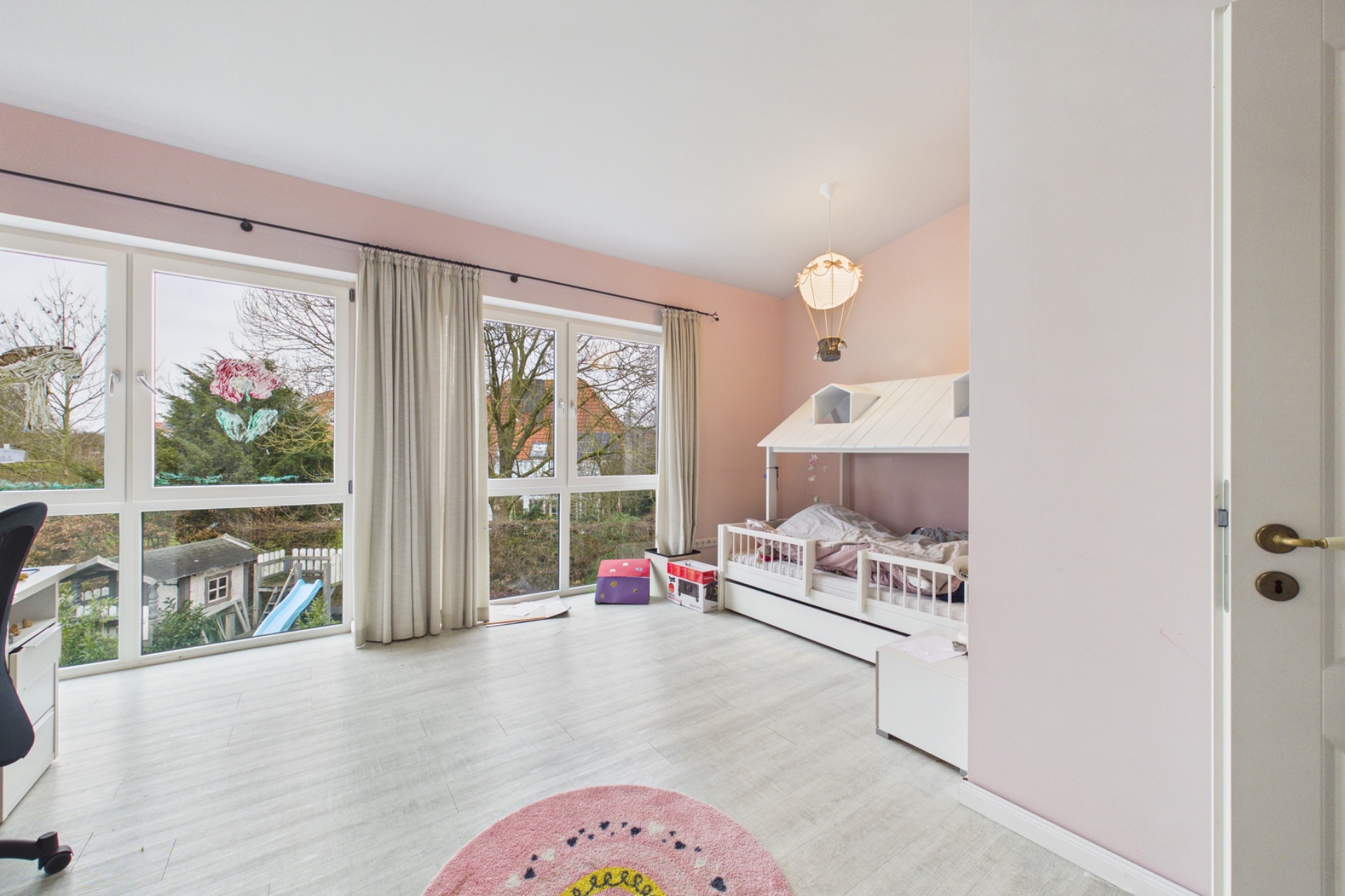 Helles Kinderzimmer mit großem Fenster, Vorhängen, Schreibtisch und einem Hausbett.