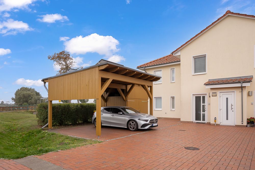 Haus mit rotem Dach und Carport, unter dem ein silbernes Auto auf gepflastertem Hof parkt.