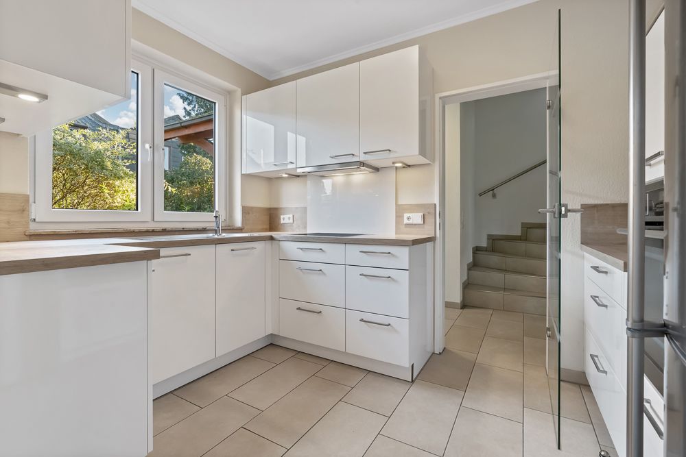 Moderne Küche mit weißen Schränken, großen Fenstern und Blick auf eine Treppe.