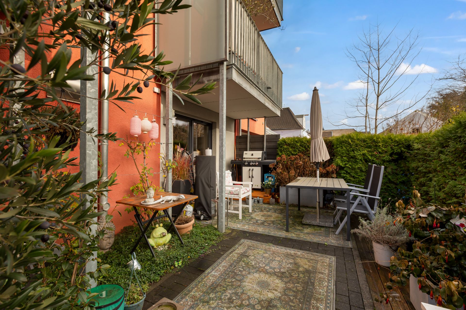 Terrasse mit Tisch, Stühlen, Pflanzen und Grill neben einem orangefarbenen Haus.