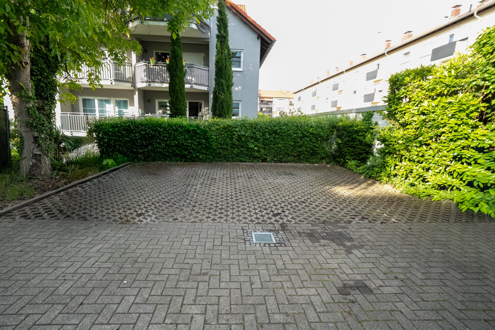 Leerer gepflasterter Parkplatz vor einem Wohnhaus, umgeben von Büschen und Bäumen.