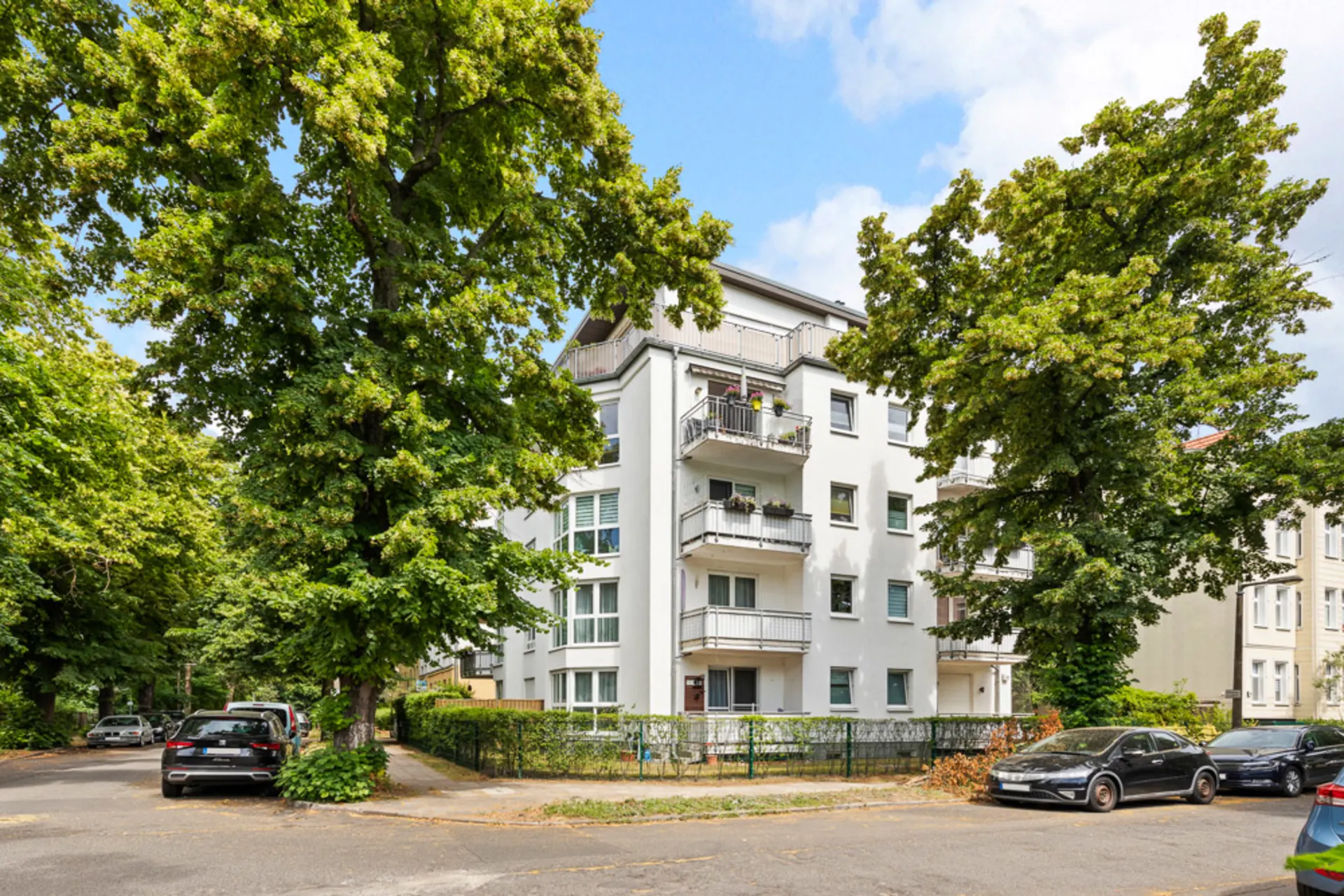 Mehrstöckiges Wohnhaus mit Balkonen, umgeben von Bäumen und geparkten Autos an der Straße.