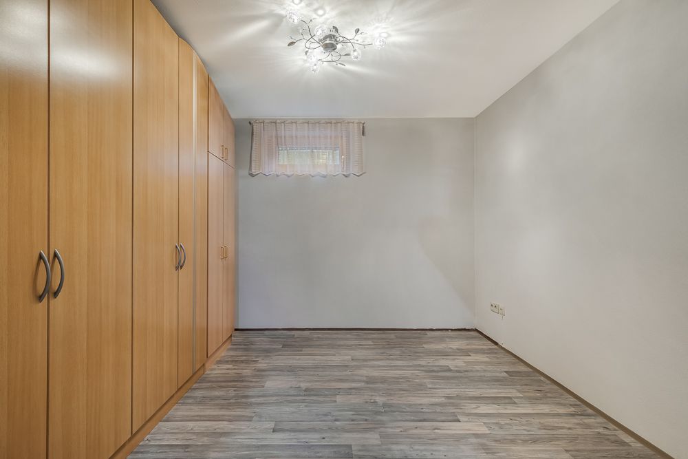 Leerer Raum mit Holzschränken, grauem Boden und Deckenleuchte, kleines Fenster mit Gardine.