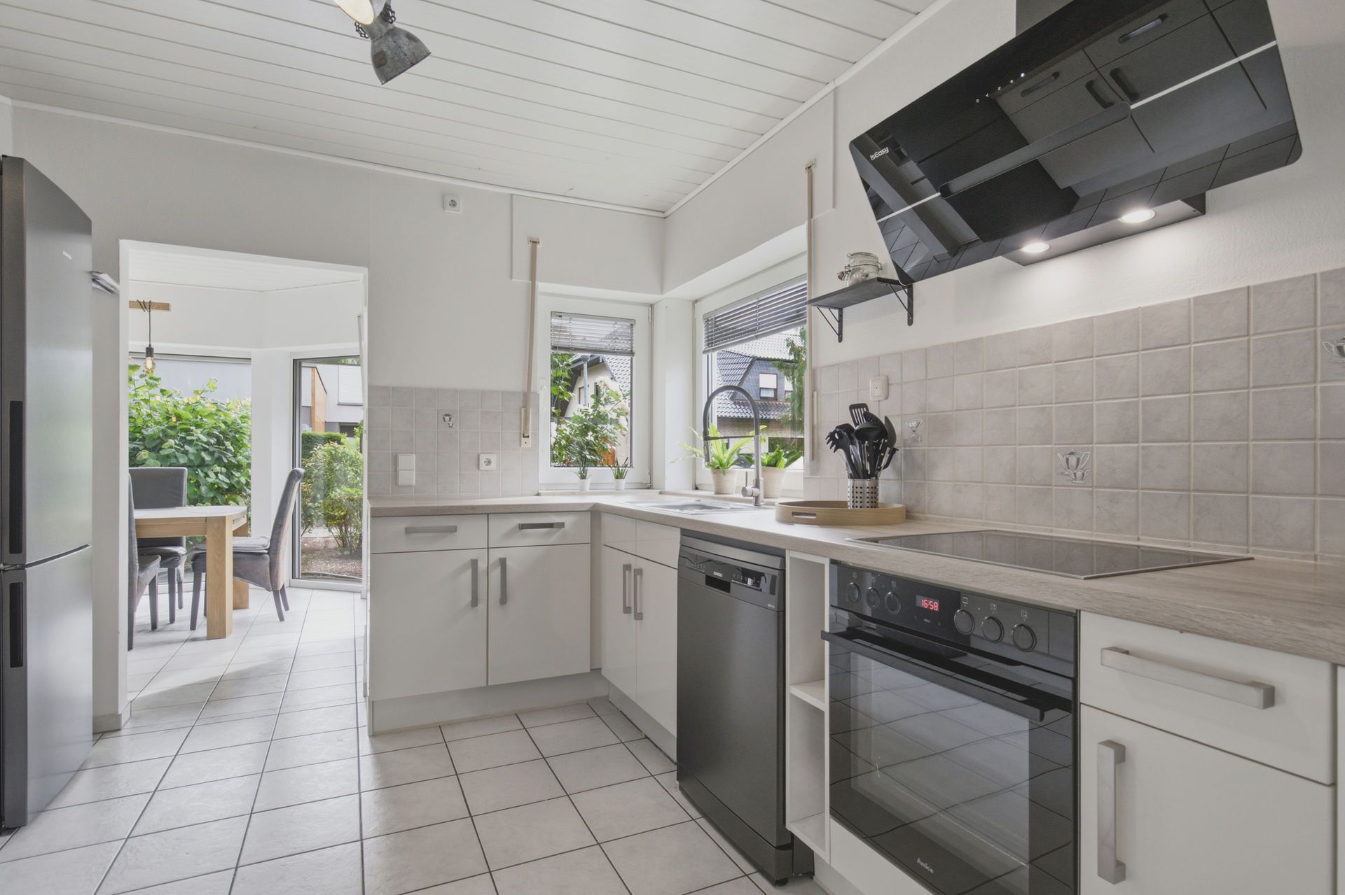 Moderne weiße Küche mit Herd, Spülmaschine und großem Fenster. Essbereich im Hintergrund sichtbar.