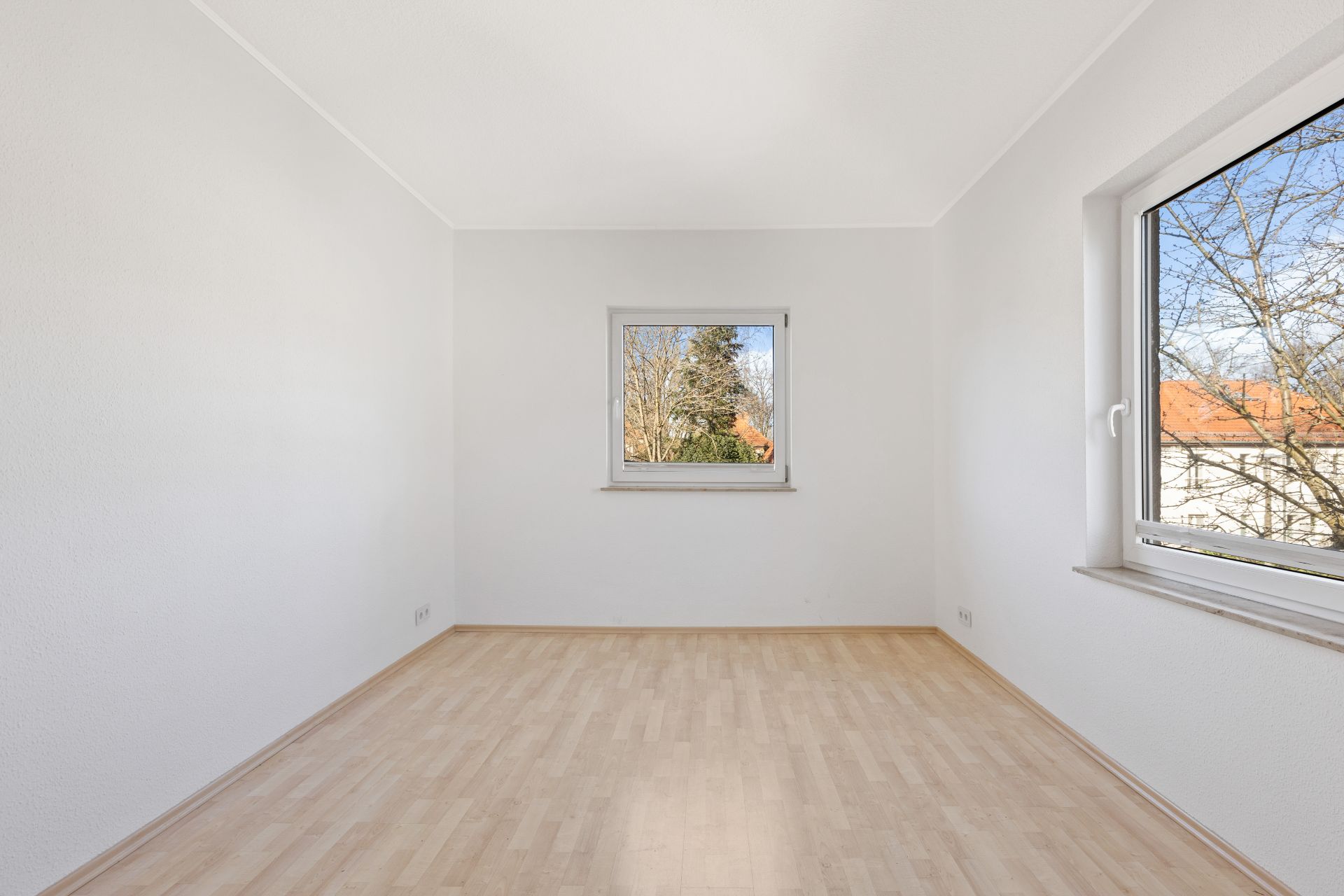 Leerer, heller Raum mit zwei Fenstern und hellem Holzboden.