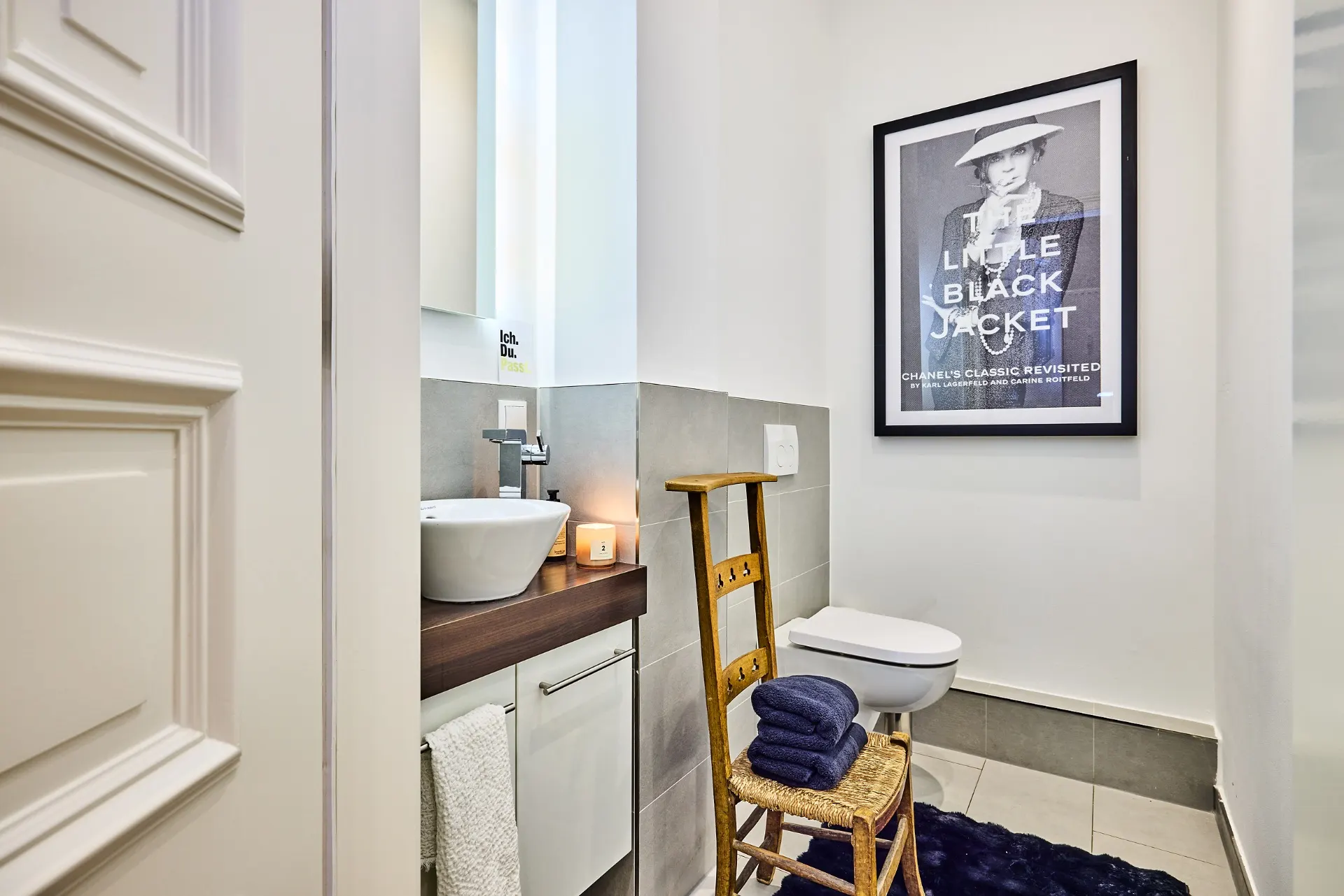 Kleines Badezimmer mit Waschbecken, Toilette, Spiegel, Stuhl mit Handtüchern und großem gerahmtem Bild an der Wand.