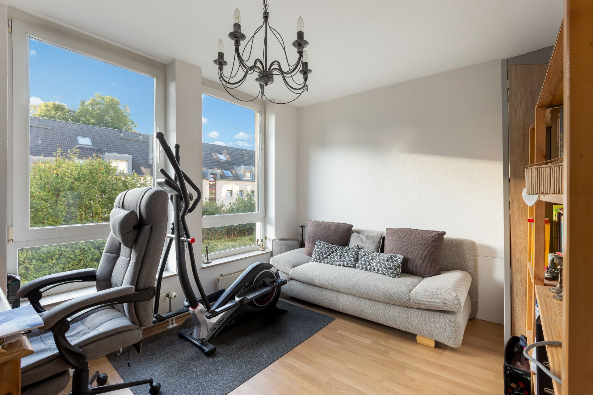 Helles Zimmer mit Sofa, Schreibtischstuhl, Heimtrainer und großem Fenster.