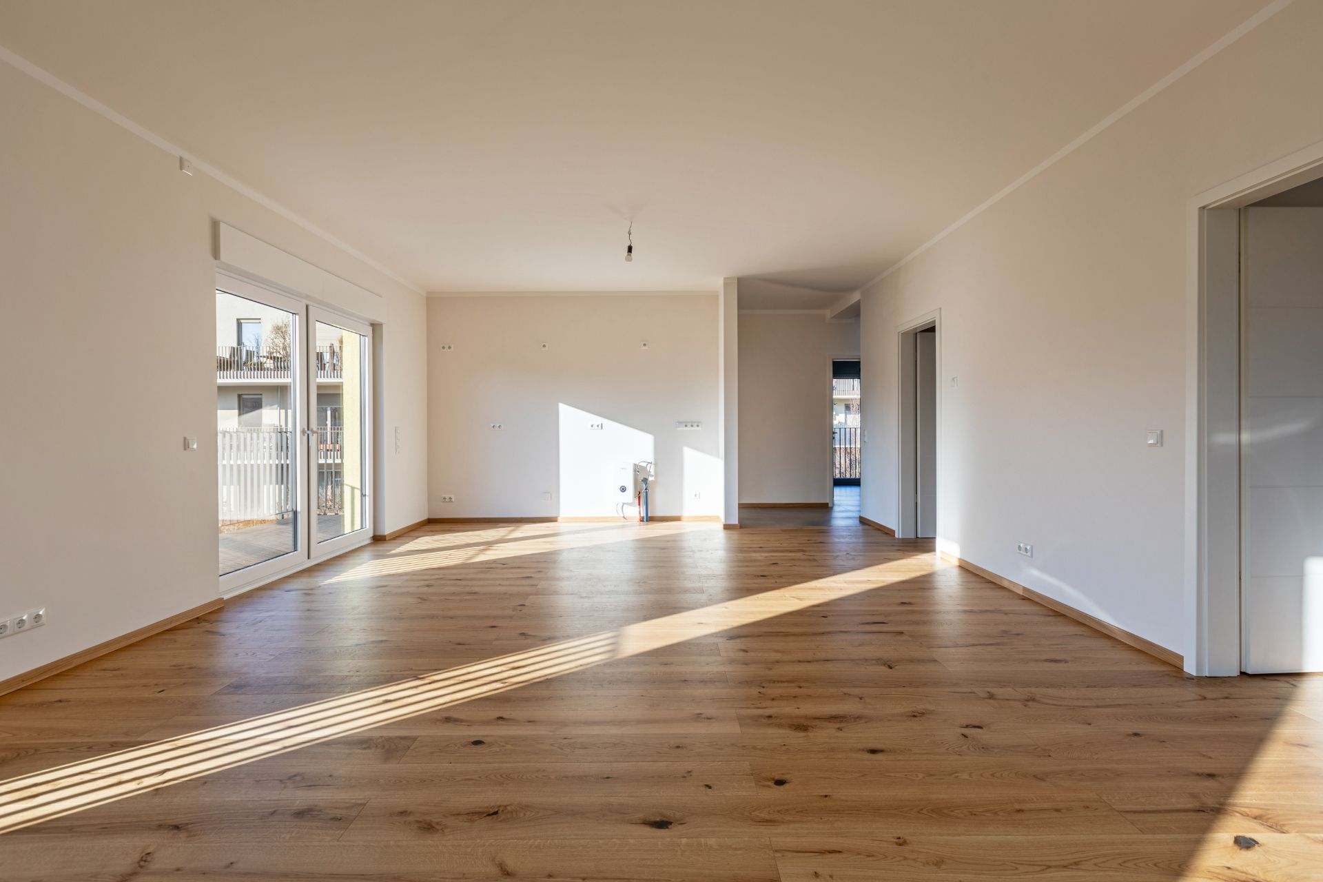Helles, leeres Wohnzimmer mit Parkettboden und großen Fenstertüren zu einem Balkon.