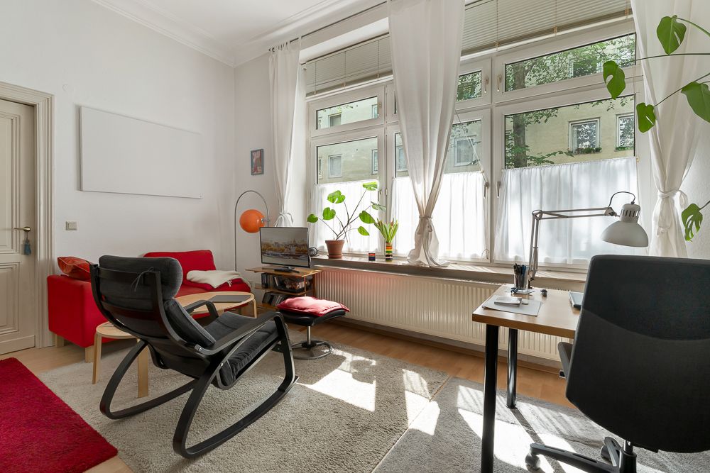 Helles Wohnzimmer mit großen Fenstern, Schreibtisch, Stuhl, Sessel und Zimmerpflanzen.