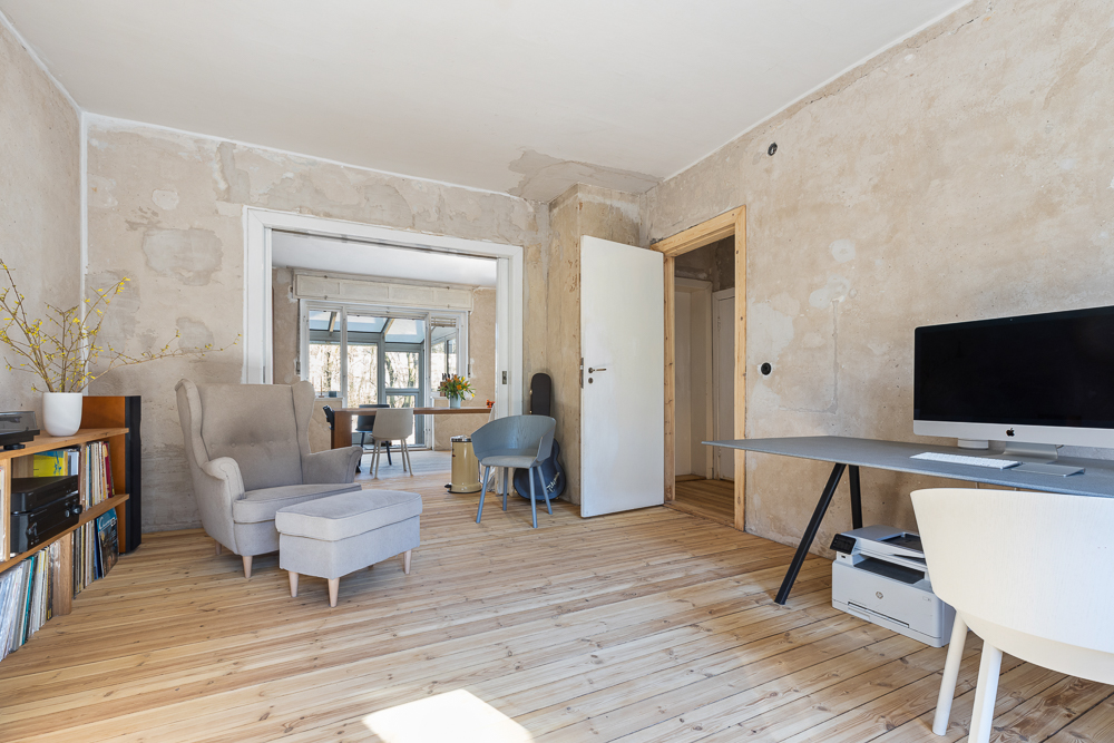 Wohnzimmer mit Sessel, Schreibtisch, Computer und Bücherregal auf hellem Holzboden.