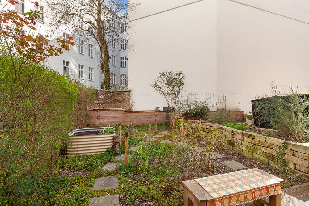 Kleiner Garten mit Steinen, Hochbeeten, Pflanzen und einem Holztisch hinter einem Wohnhaus.