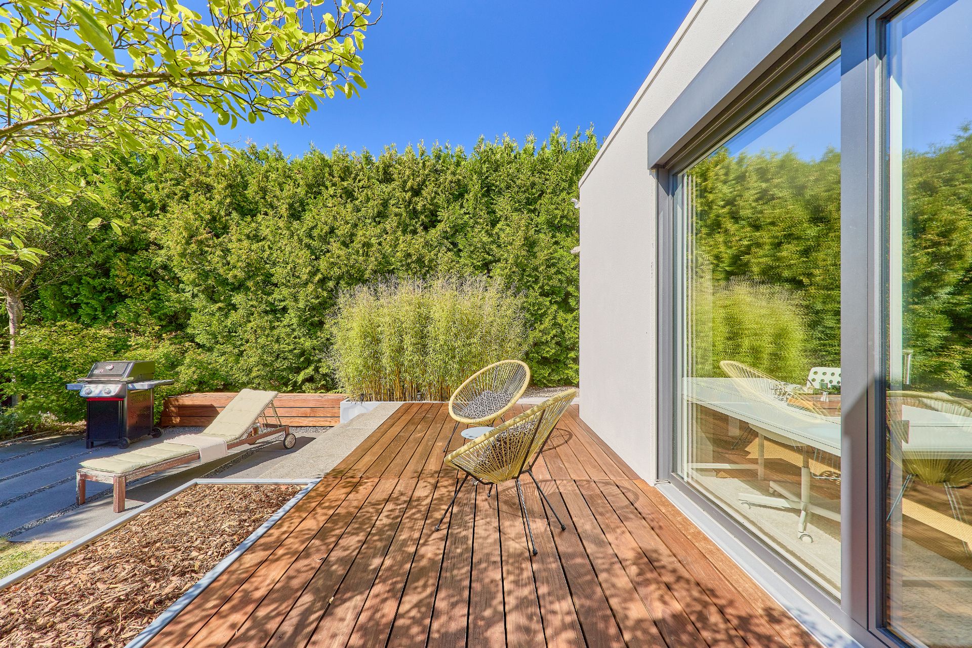 Holzterrasse mit zwei Stühlen, Liege, Grill und Blick auf grüne Hecke bei sonnigem Wetter.