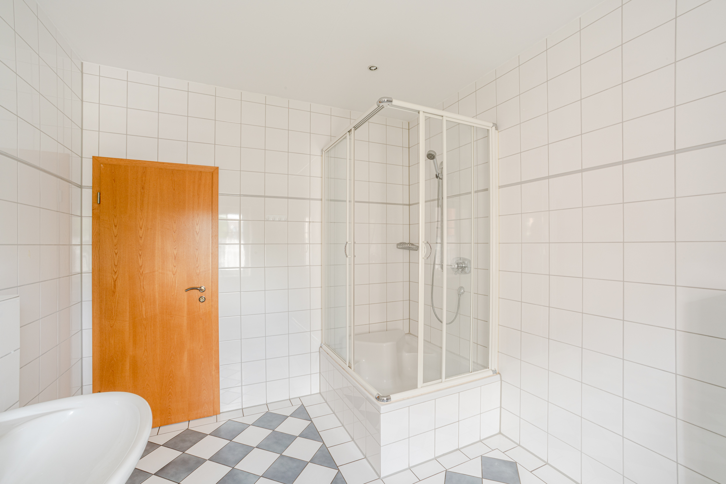 Helles Bad mit Eckdusche, Holztür und weißen Fliesen an Wand und Boden.