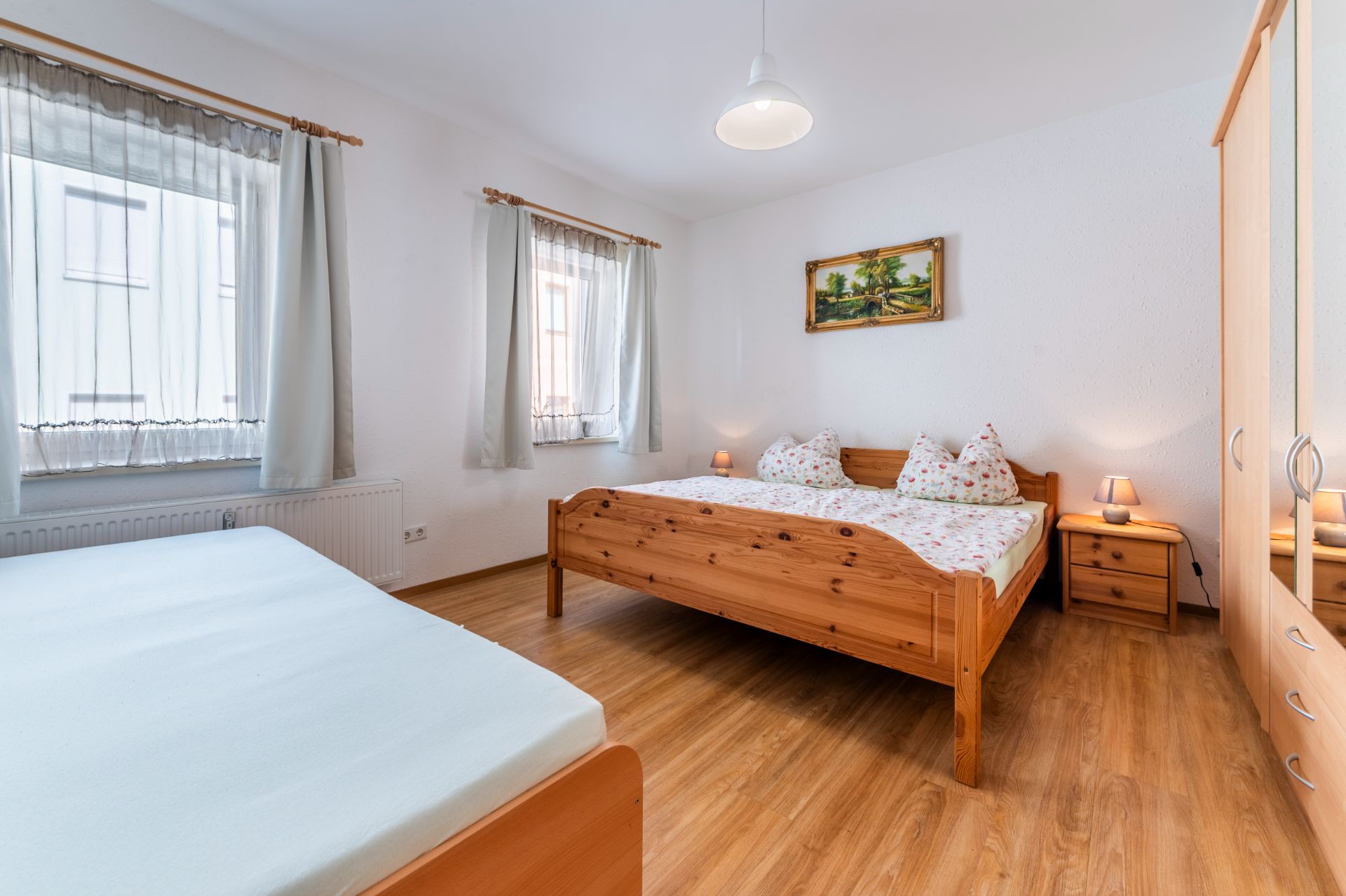 Helles Schlafzimmer mit Holzboden, Doppelbett, Einzelbett, Kleiderschrank und zwei Fenstern.