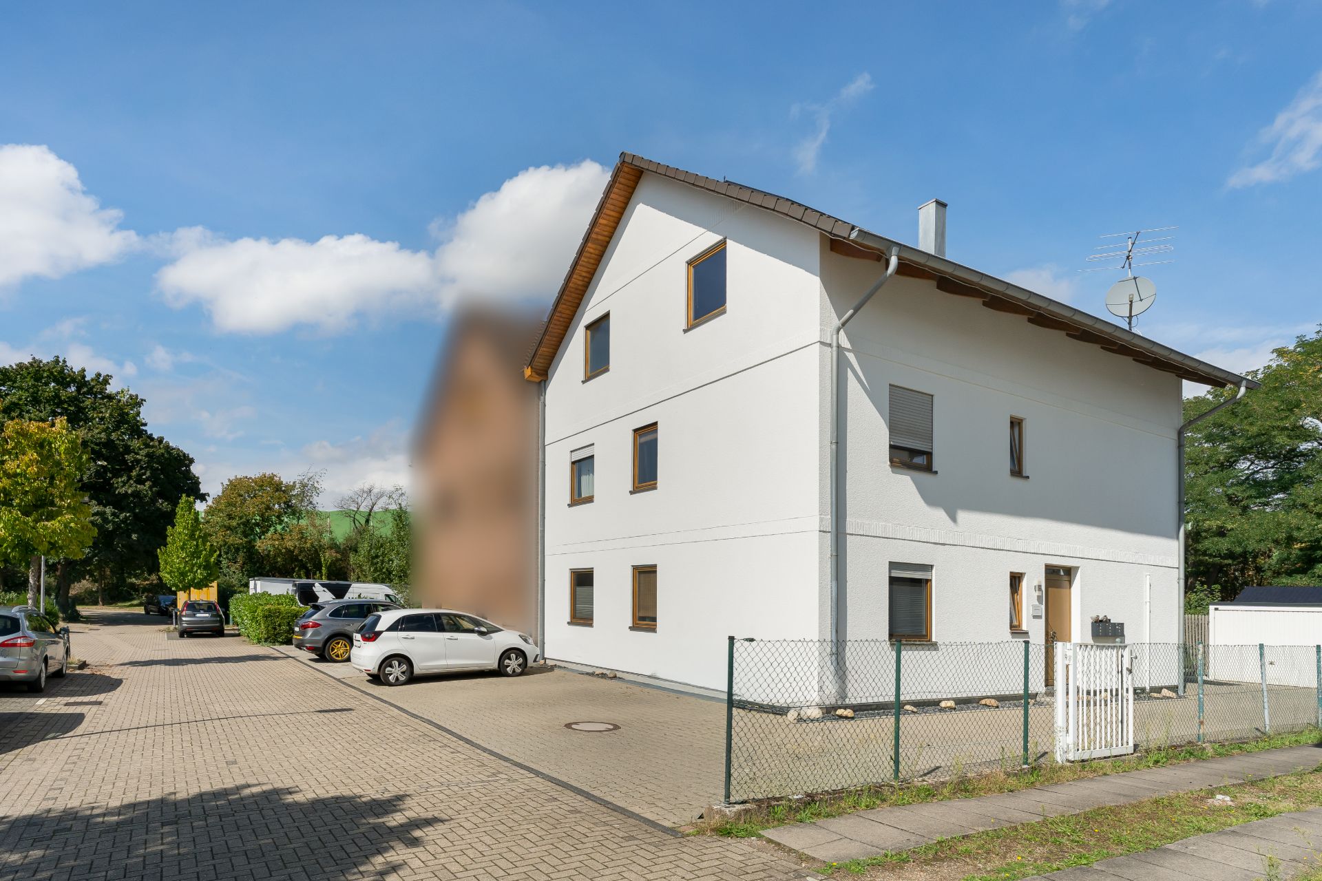 Modernes, weißes Wohnhaus mit drei Etagen, umzäuntem Garten und parkenden Autos davor.