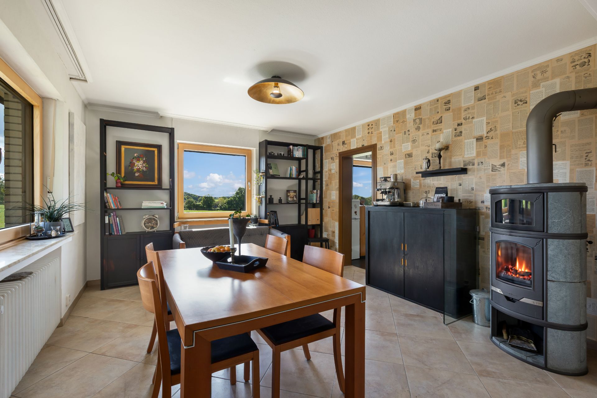 Esszimmer mit Holztisch, vier Stühlen, Kaminofen, Regalen und großen Fenstern mit Ausblick.