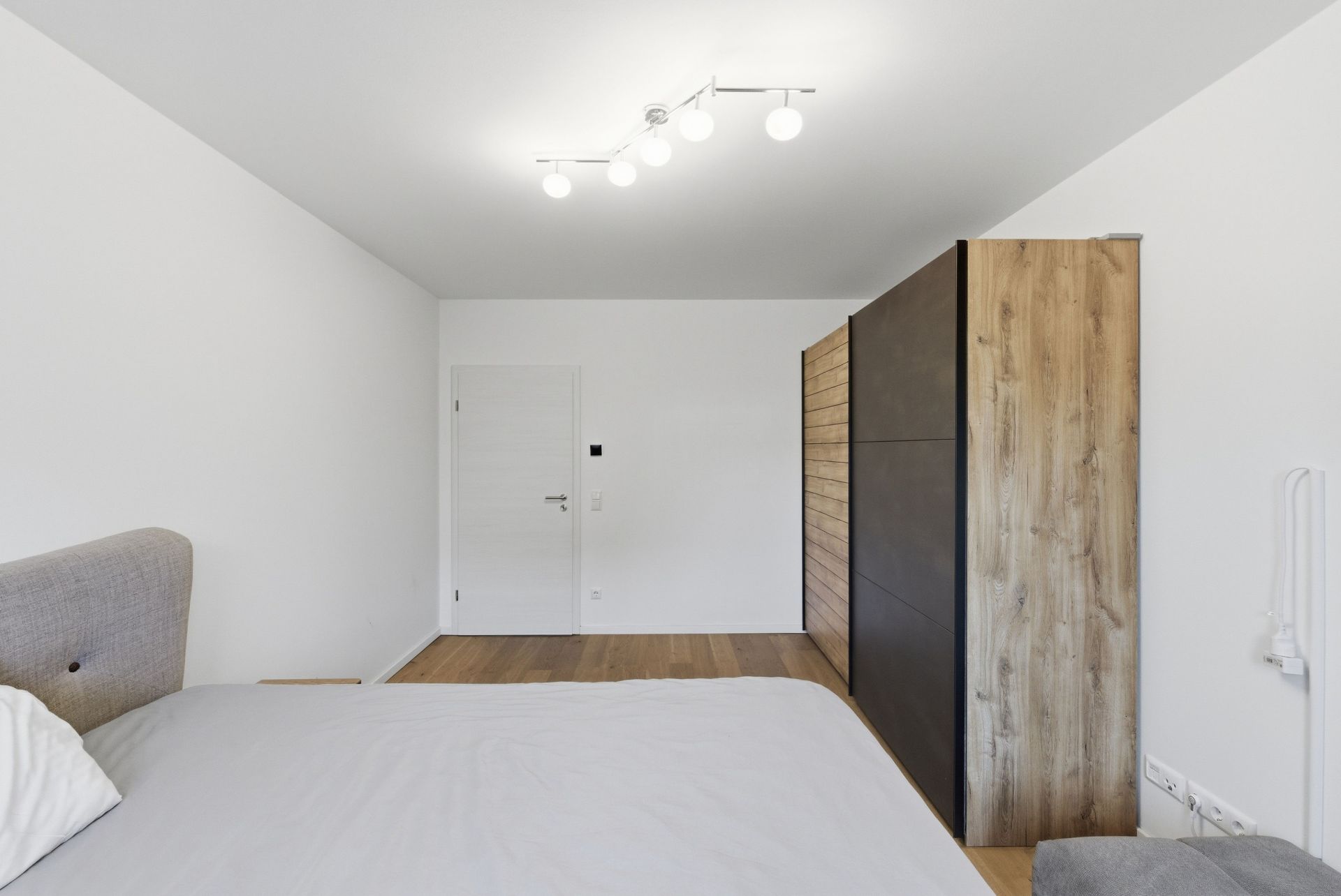 Helles Schlafzimmer mit Bett, großem Holzschrank und weißer Tür auf Holzboden.