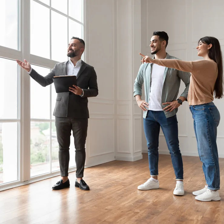 Drei Personen stehen in einem hellen Raum mit großen Fenstern und Holzfußboden.