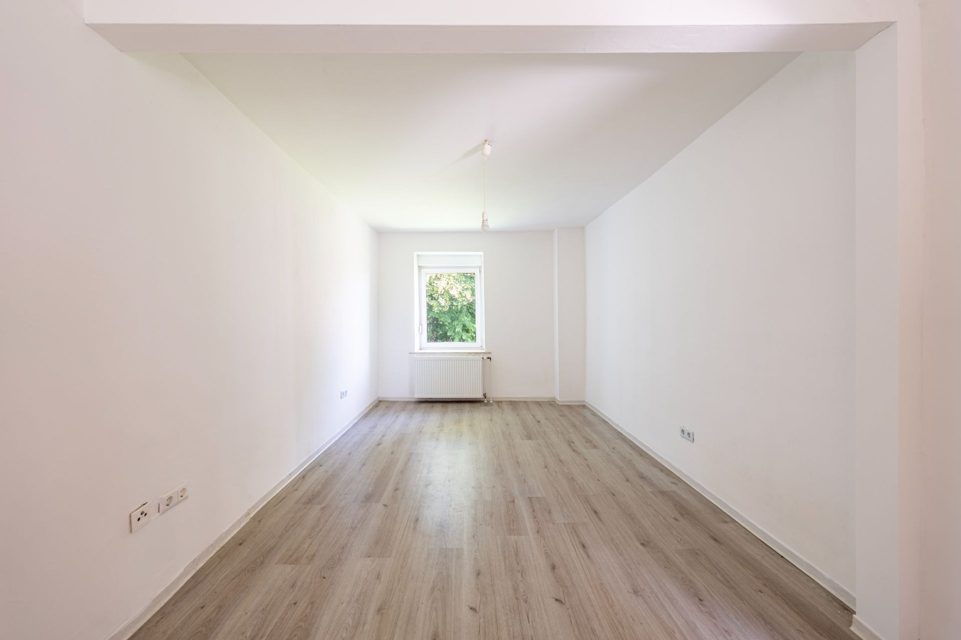 Leerer Raum mit hellem Holzfußboden, weißen Wänden und einem Fenster am Ende.