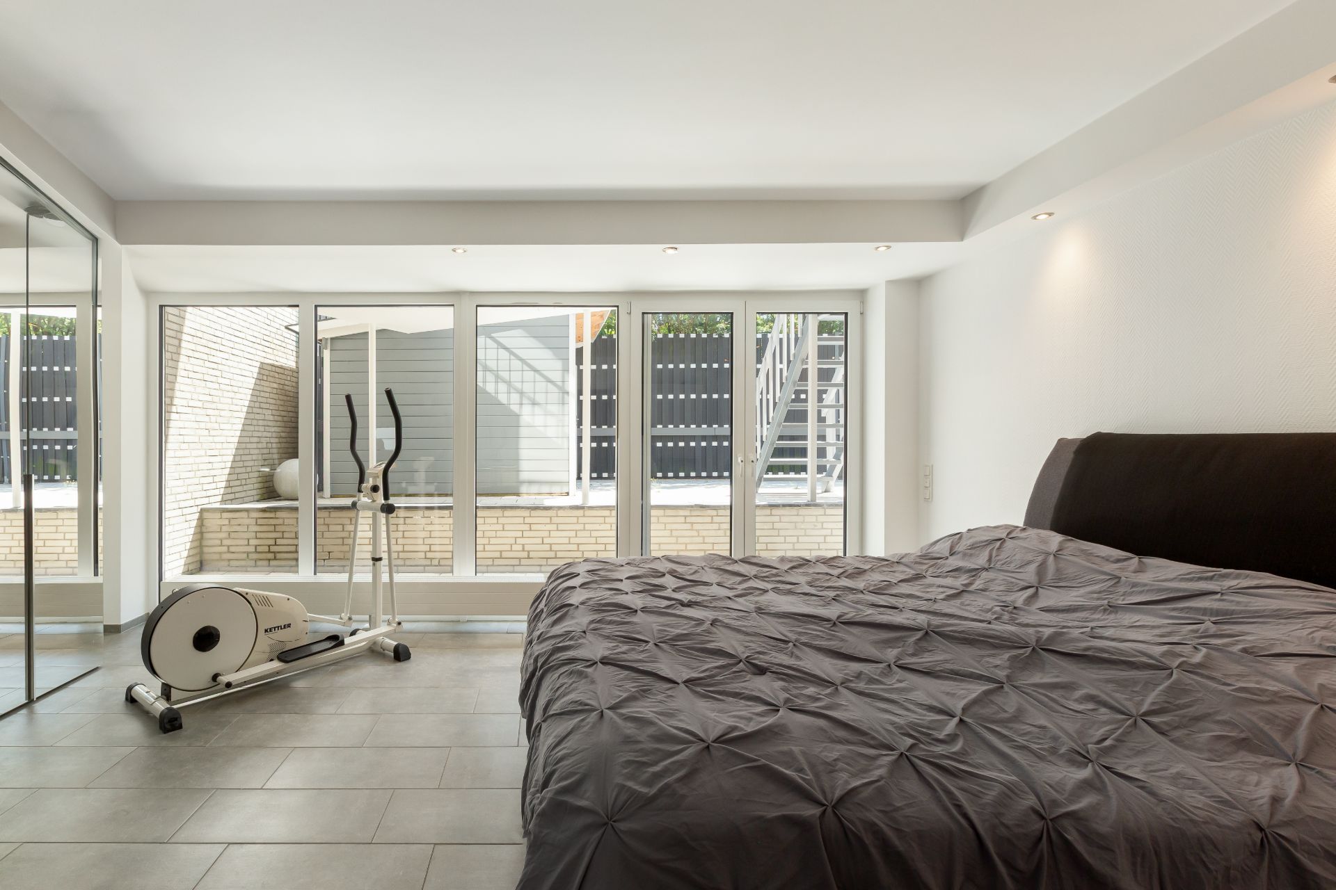 Schlafzimmer mit großem Bett, Ellipsentrainer und bodentiefen Fenstern mit Blick auf Terrasse.