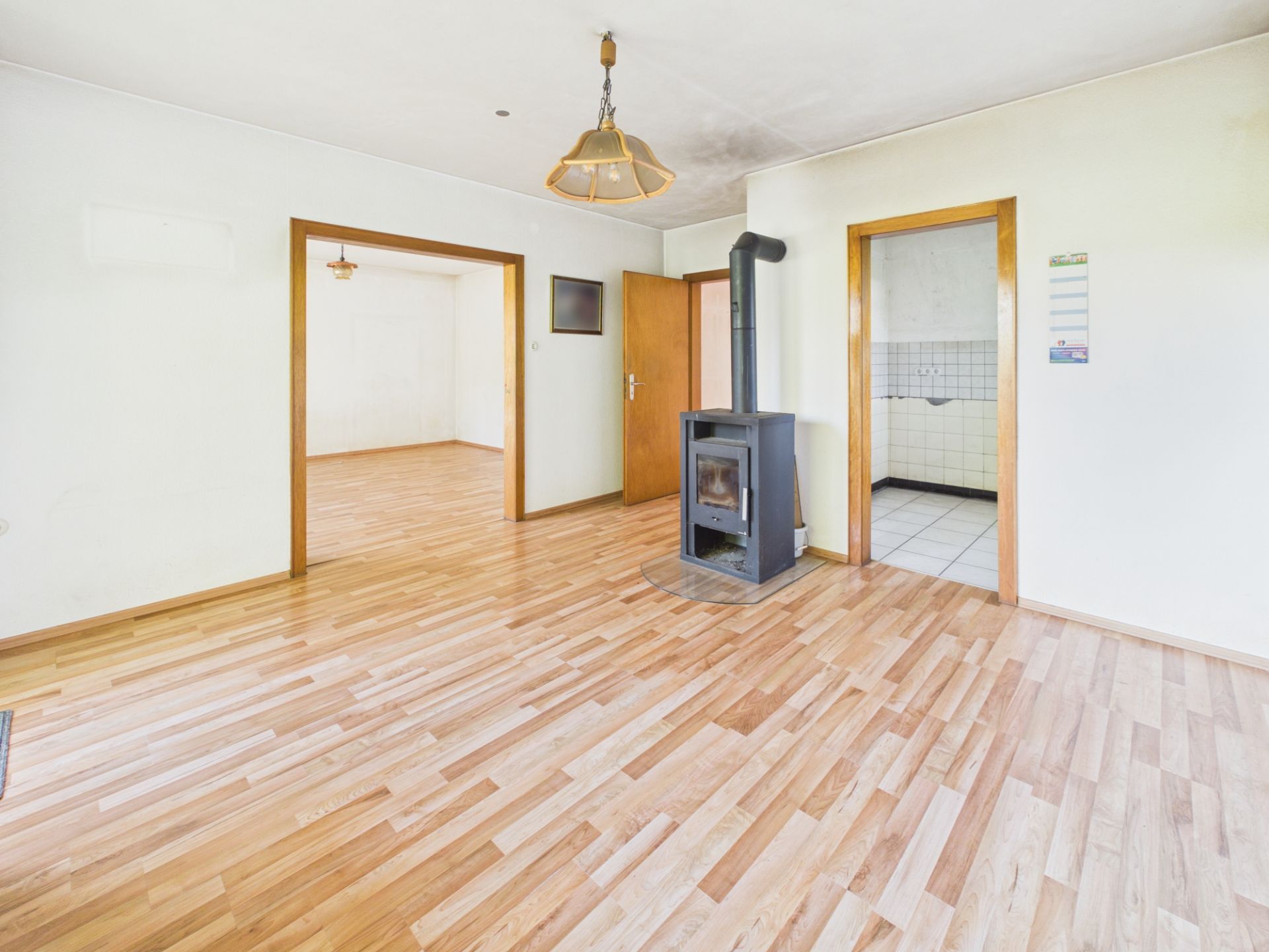 Helles Wohnzimmer mit Holzfußboden, Kaminofen und Zugang zur Küche und einem weiteren Raum.