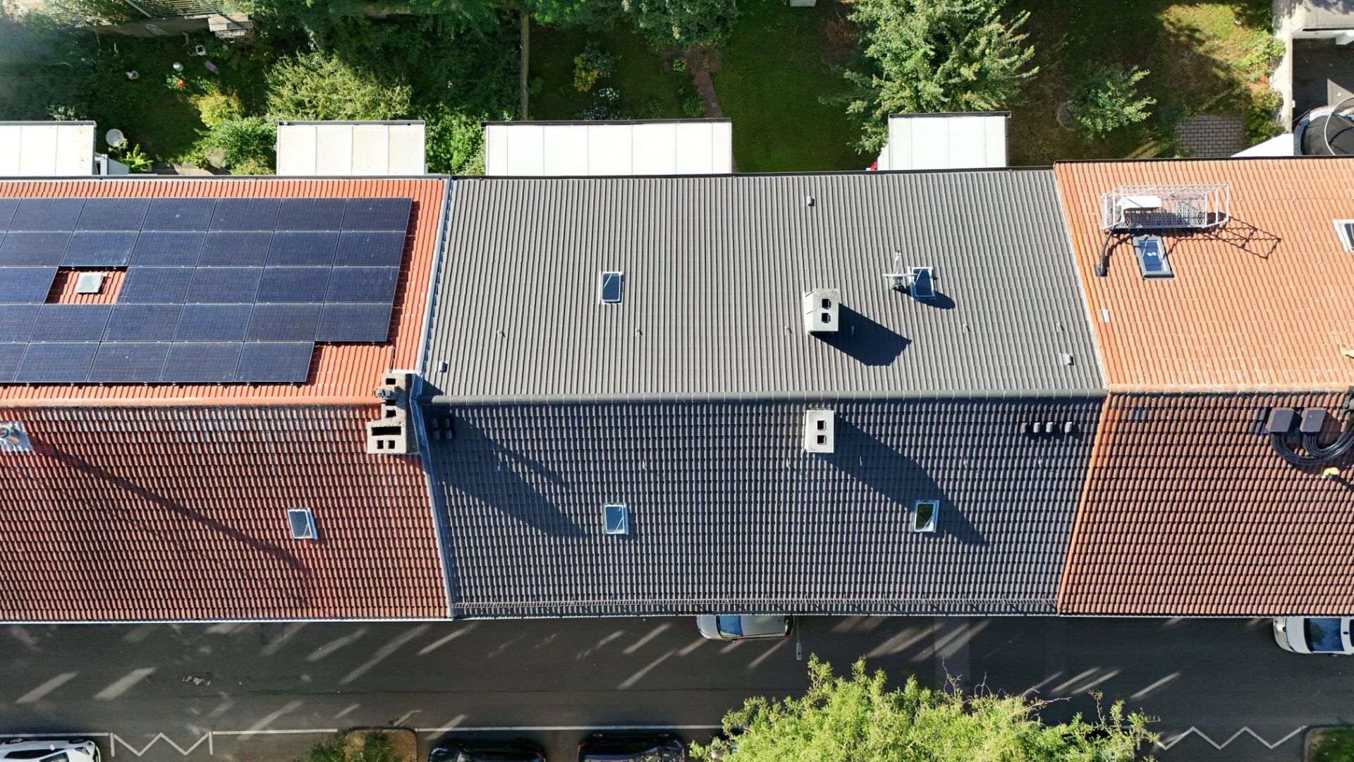 Draufsicht auf drei Hausdächer, eines davon mit Solarpanels, daneben graue und rote Dächer.