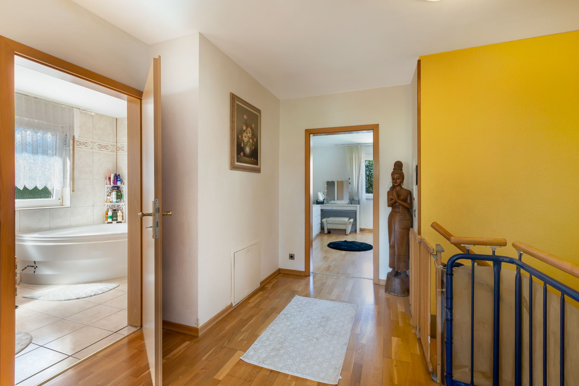 Heller Flur mit Holzfußboden, Blick ins Badezimmer und Schlafzimmer, gelbe Wand und Statue neben Treppe.