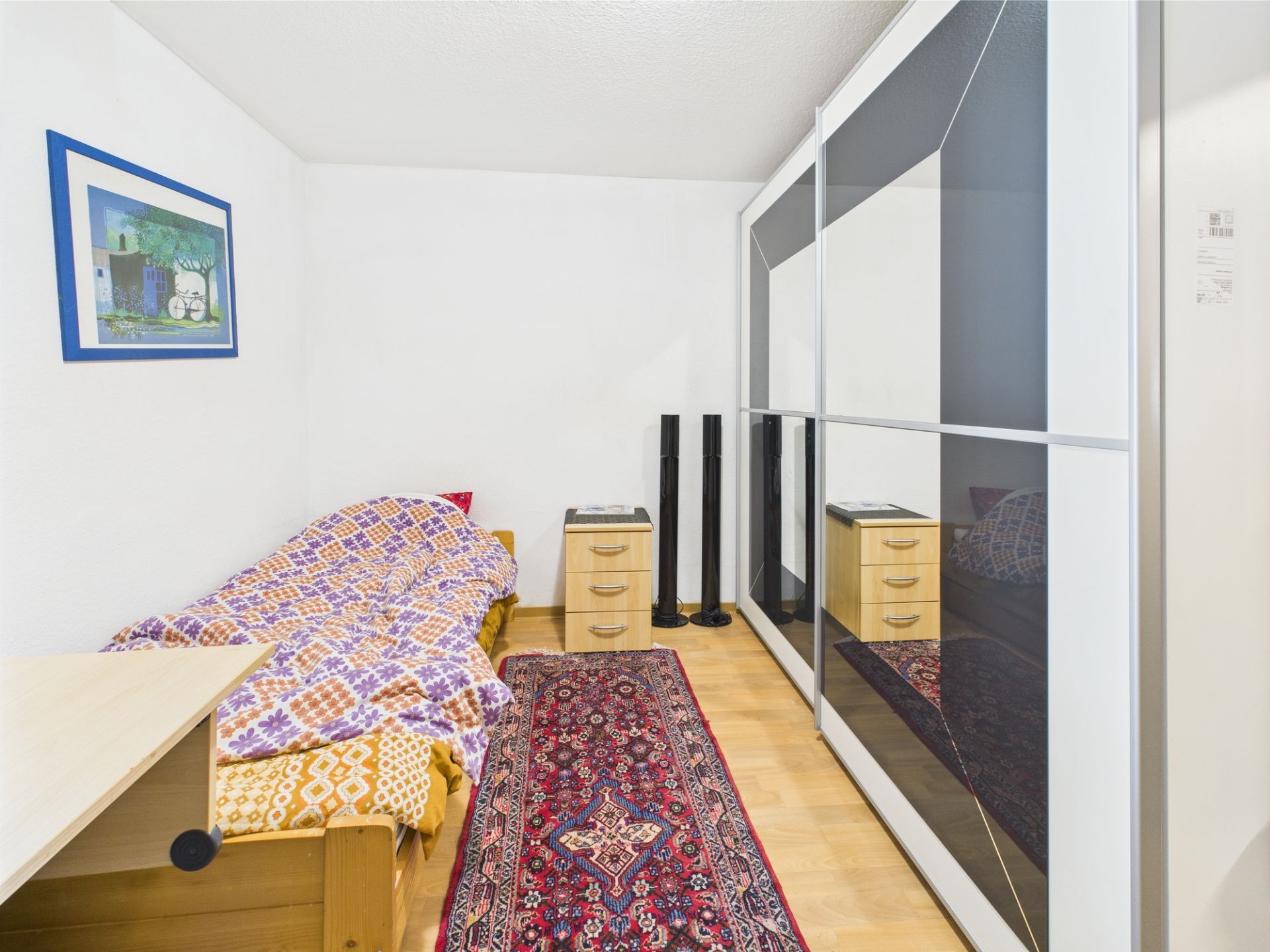 Schlafzimmer mit Einzelbett, großem Schrank, Nachttisch, Teppich und Wandbild.