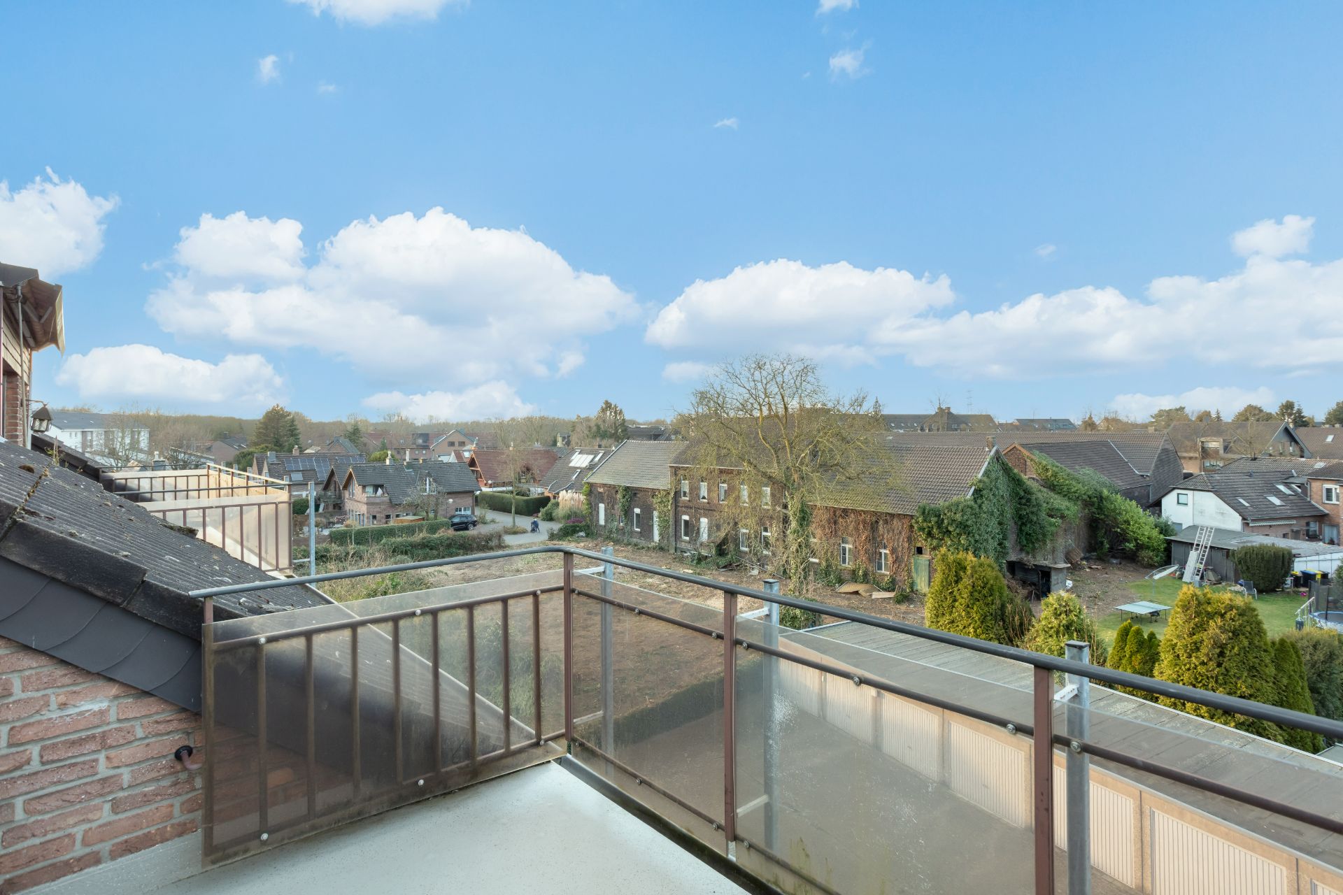 Aussicht von einem Balkon auf Nachbarhäuser, Bäume und einen blauen Himmel mit Wolken.