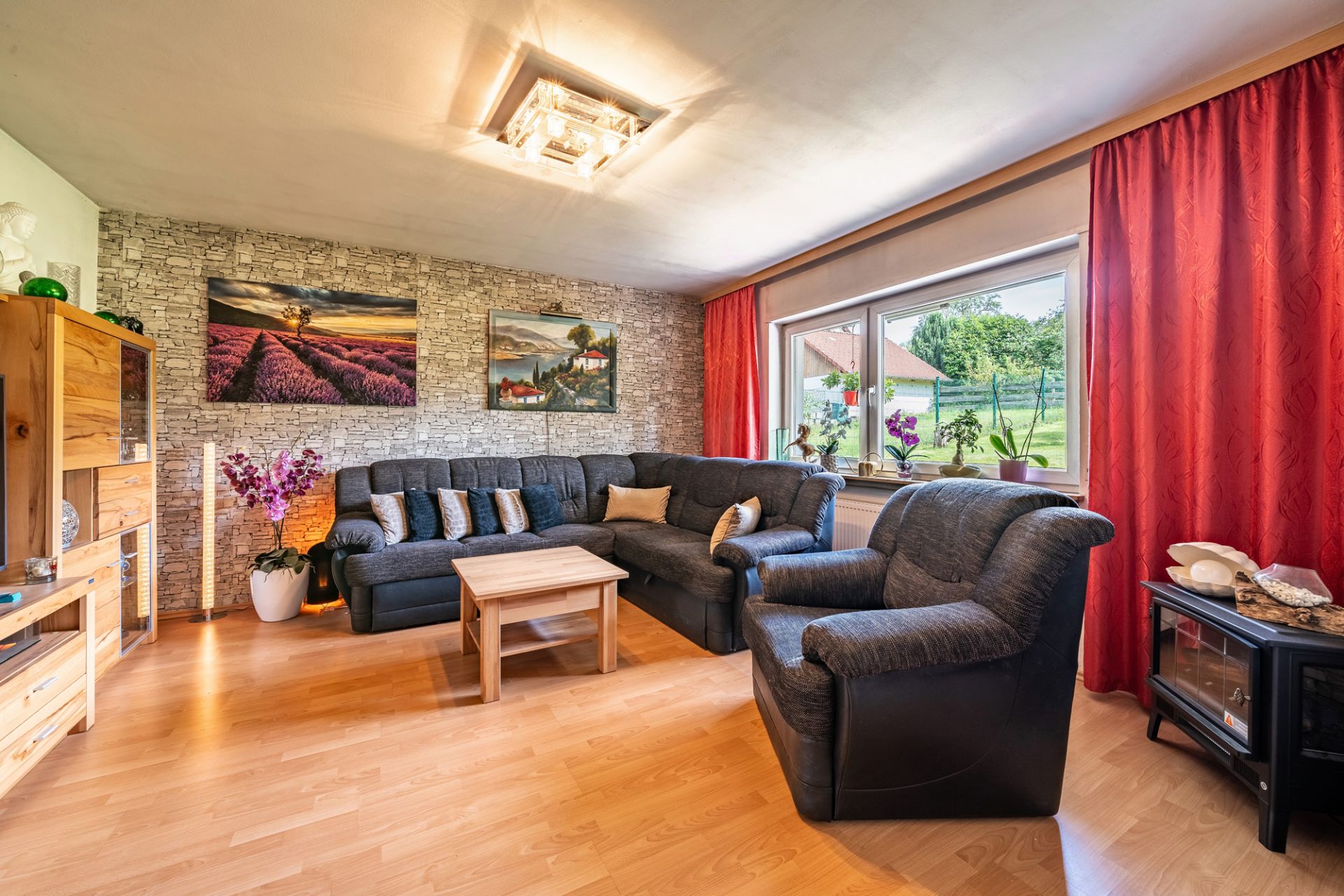 Helles Wohnzimmer mit Ecksofa, Sessel, Couchtisch, Holzmöbeln und großem Fenster mit roten Vorhängen.