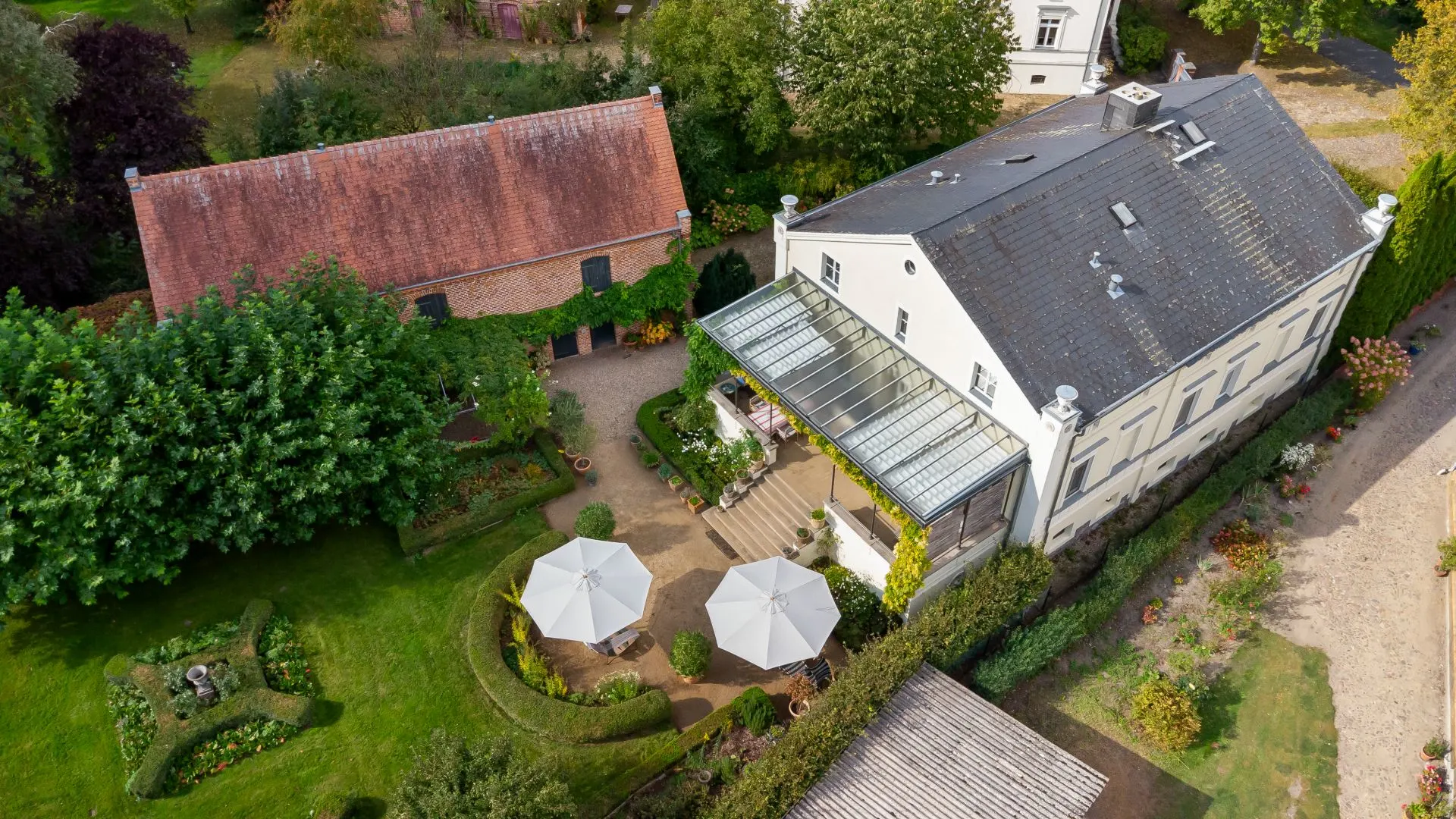 Luftaufnahme eines Wohnhauses mit Garten, Terrasse, zwei weißen Sonnenschirmen und Nebengebäude.