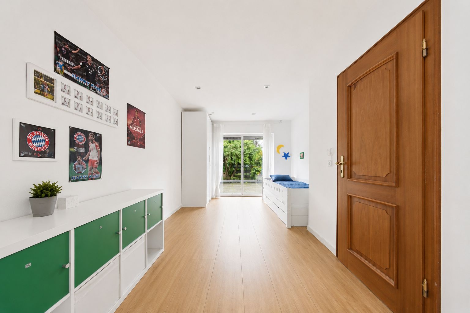 Helles Kinderzimmer mit Holzboden, grünen Regalen, Bett und Zugang zum Garten.