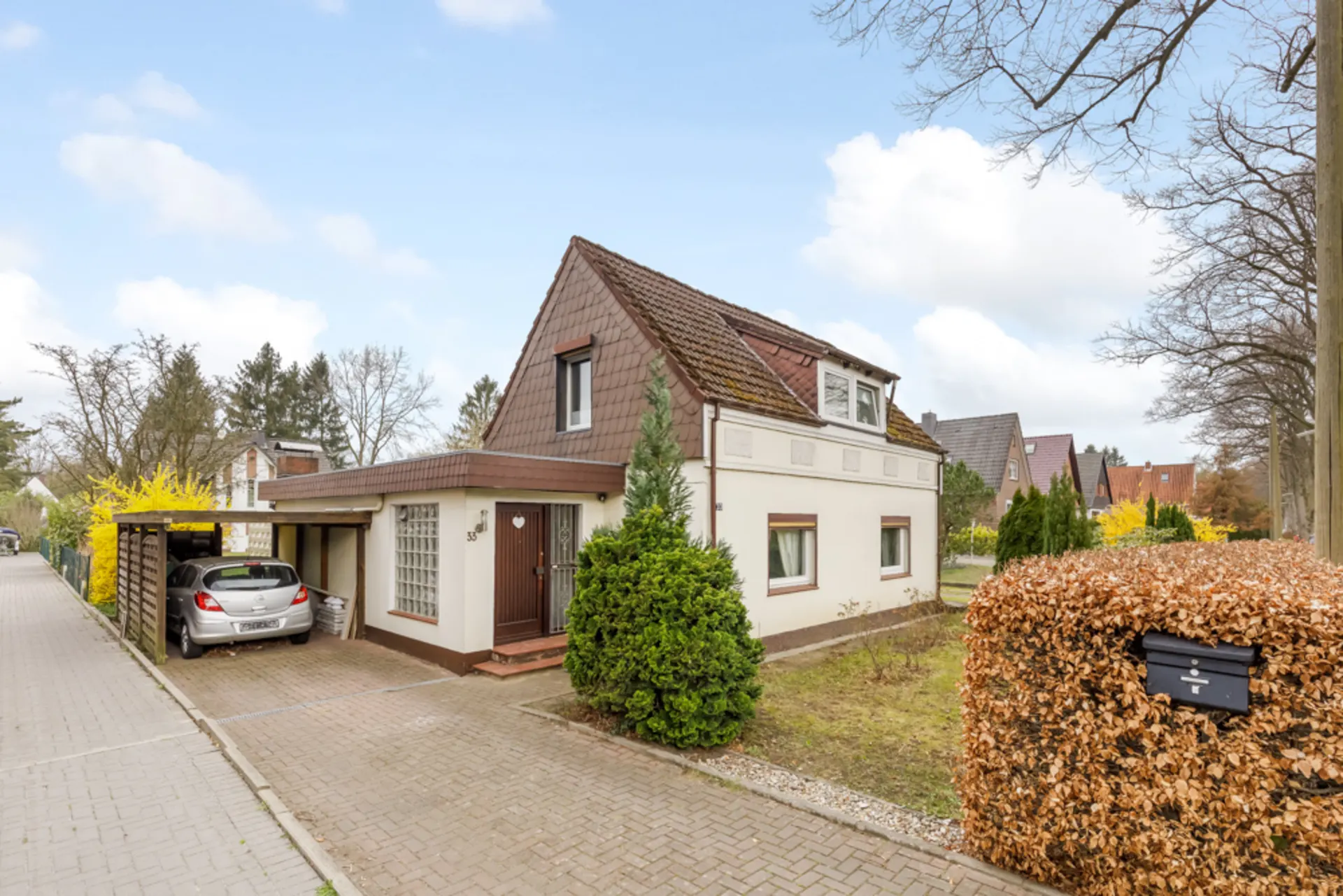 Einfamilienhaus mit Satteldach, Carport und Auto daneben, umgeben von Bäumen und Hecken.